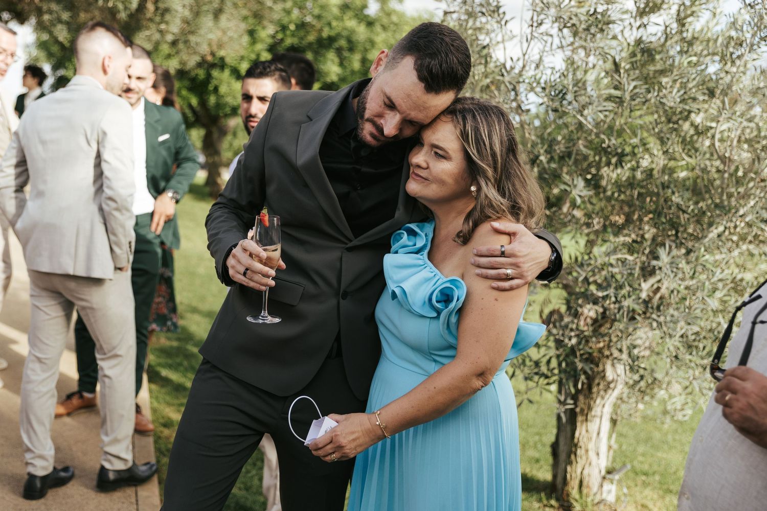 A couple embraces at an outdoor reception while other guests mingle in the background.