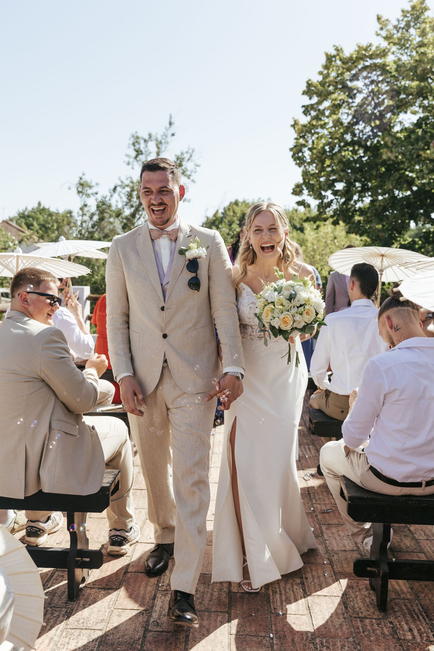 A joyful newlywed couple walks down the aisle at an outdoor wedding ceremony on a sunny day with guests seated nearby.