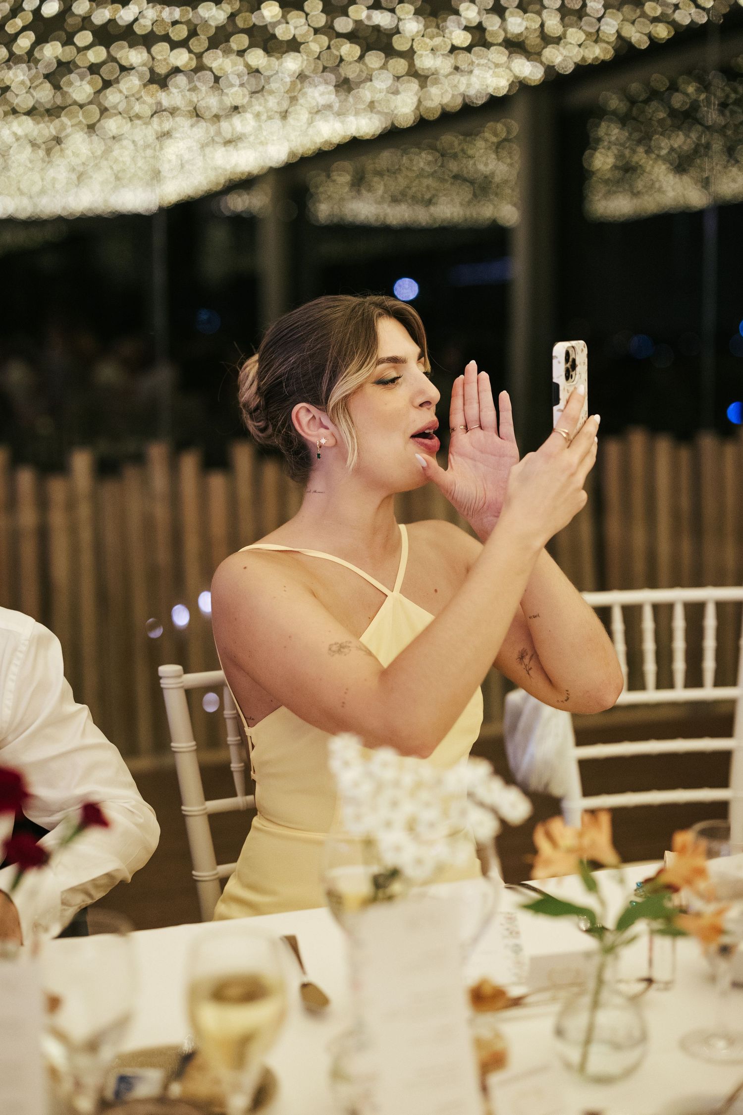 A guest in a yellow dress takes a photo with her phone under twinkling lights at a formal reception.