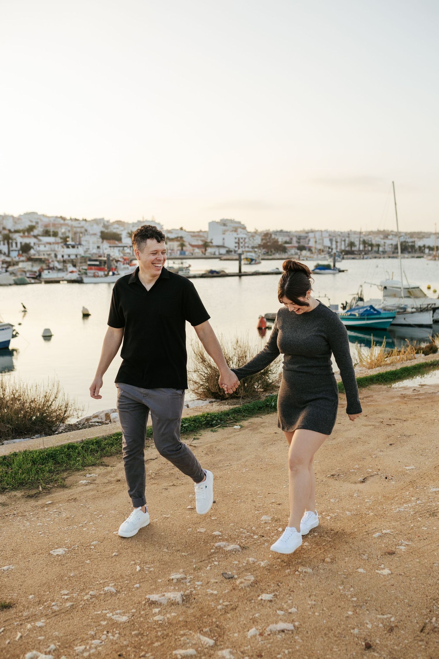 A couple walks hand in hand along a coastal path overlooking a harbor filled with boats at sunset.