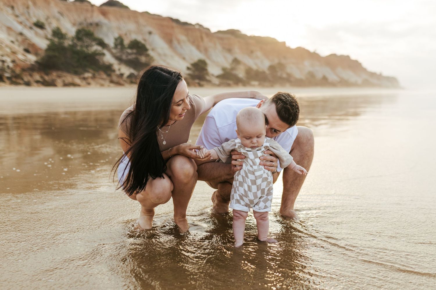 Family explores shallow beach waters together at sunset with dramatic cliffs in the background.
