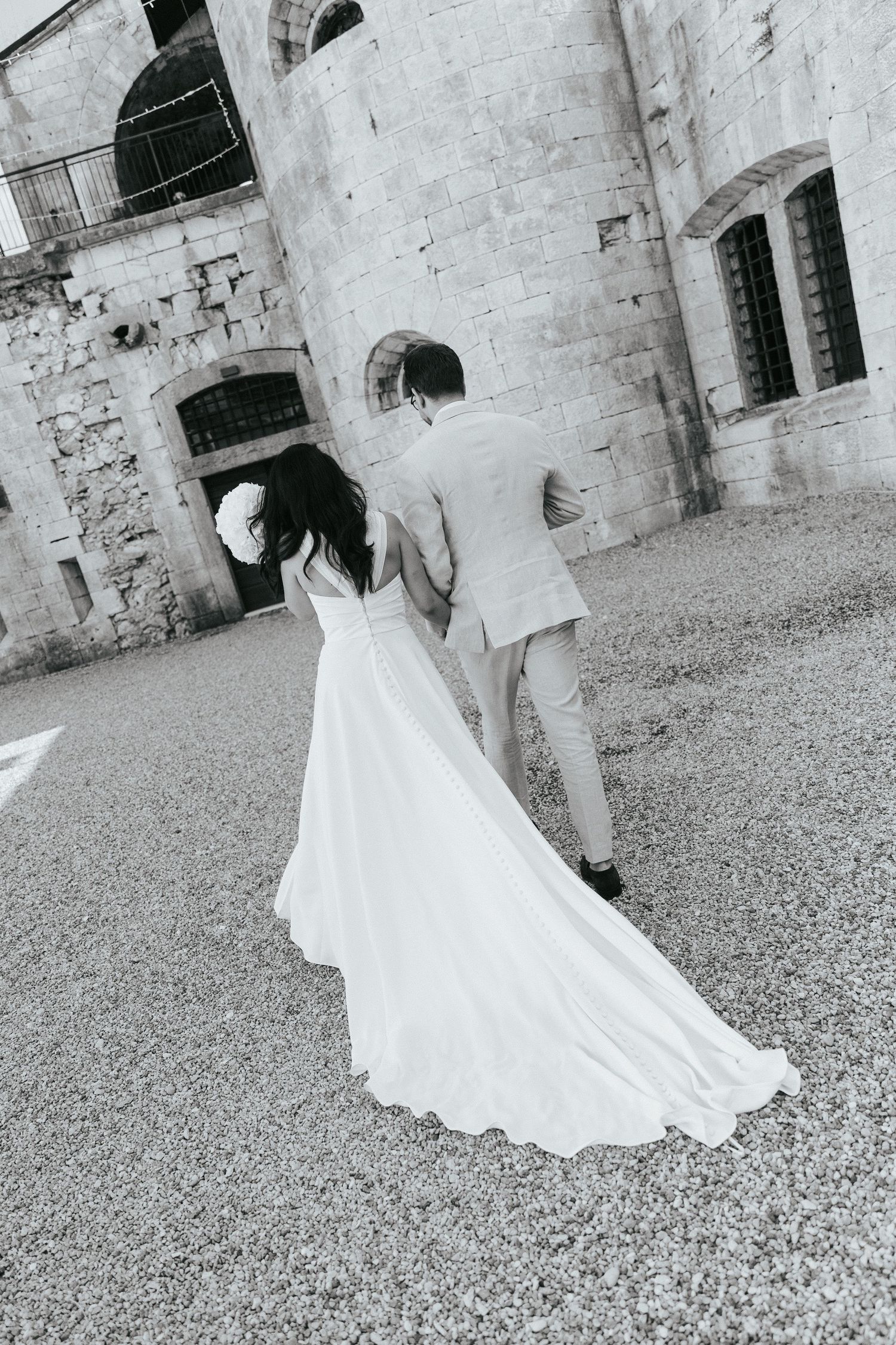 Elegant couple walking together on stone path with flowing white wedding dress trailing behind.