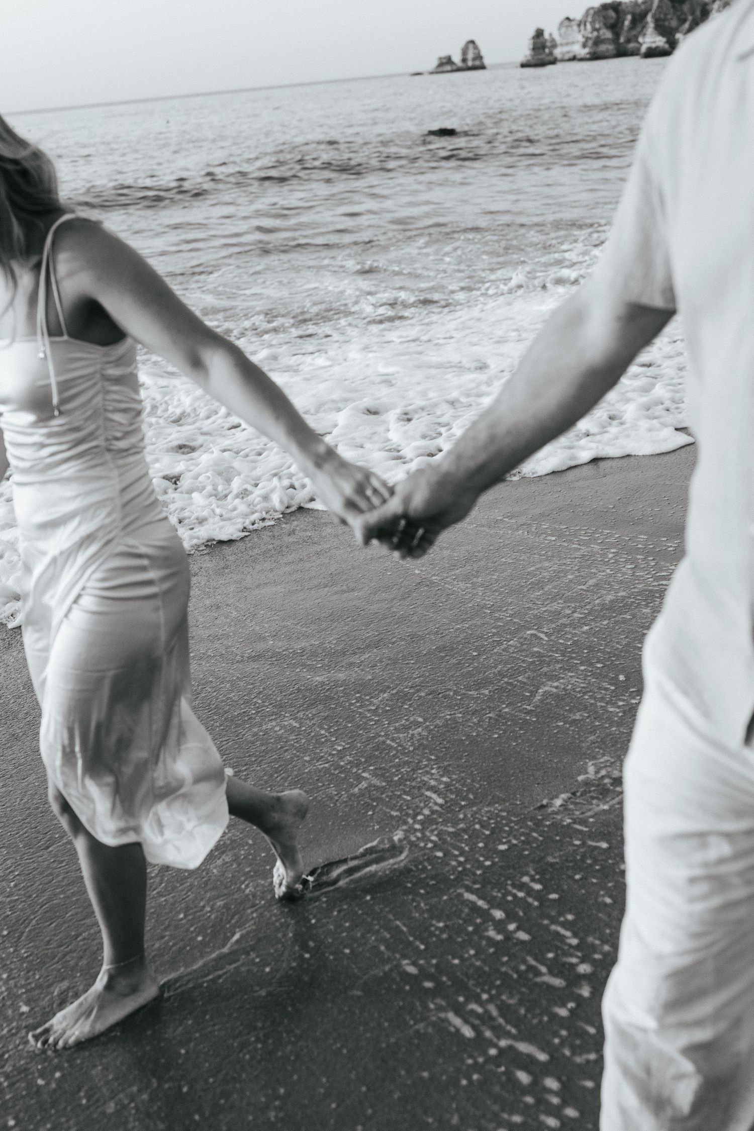 A black and white photograph shows people walking barefoot along the shoreline at the beach.