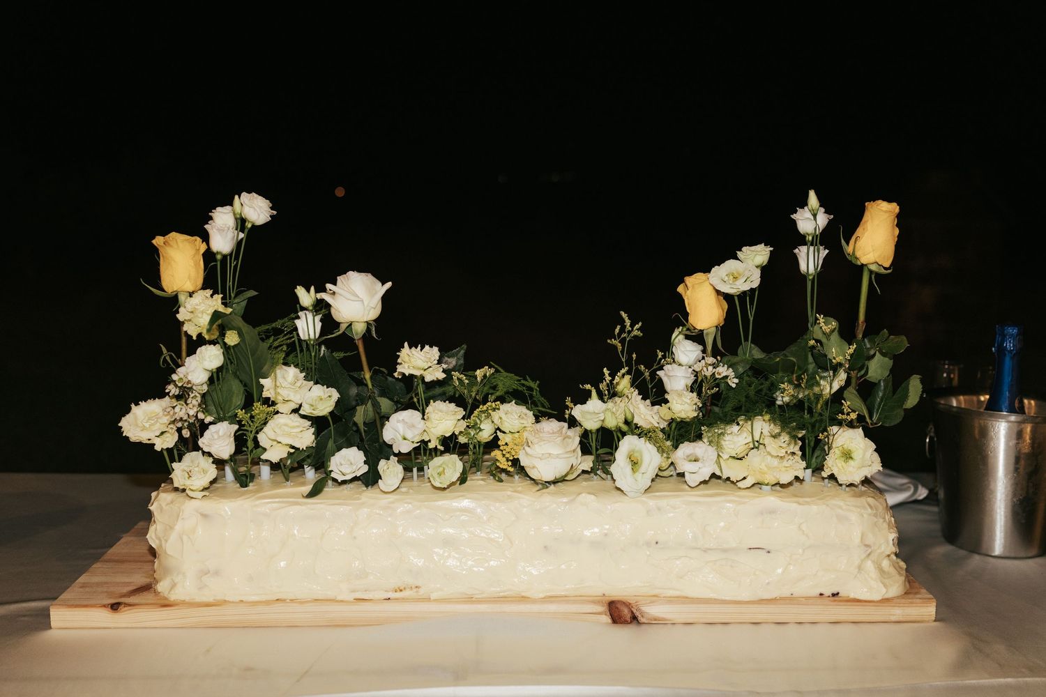 Rectangular white wedding cake decorated with yellow roses and white flowers on wooden board against dark background.