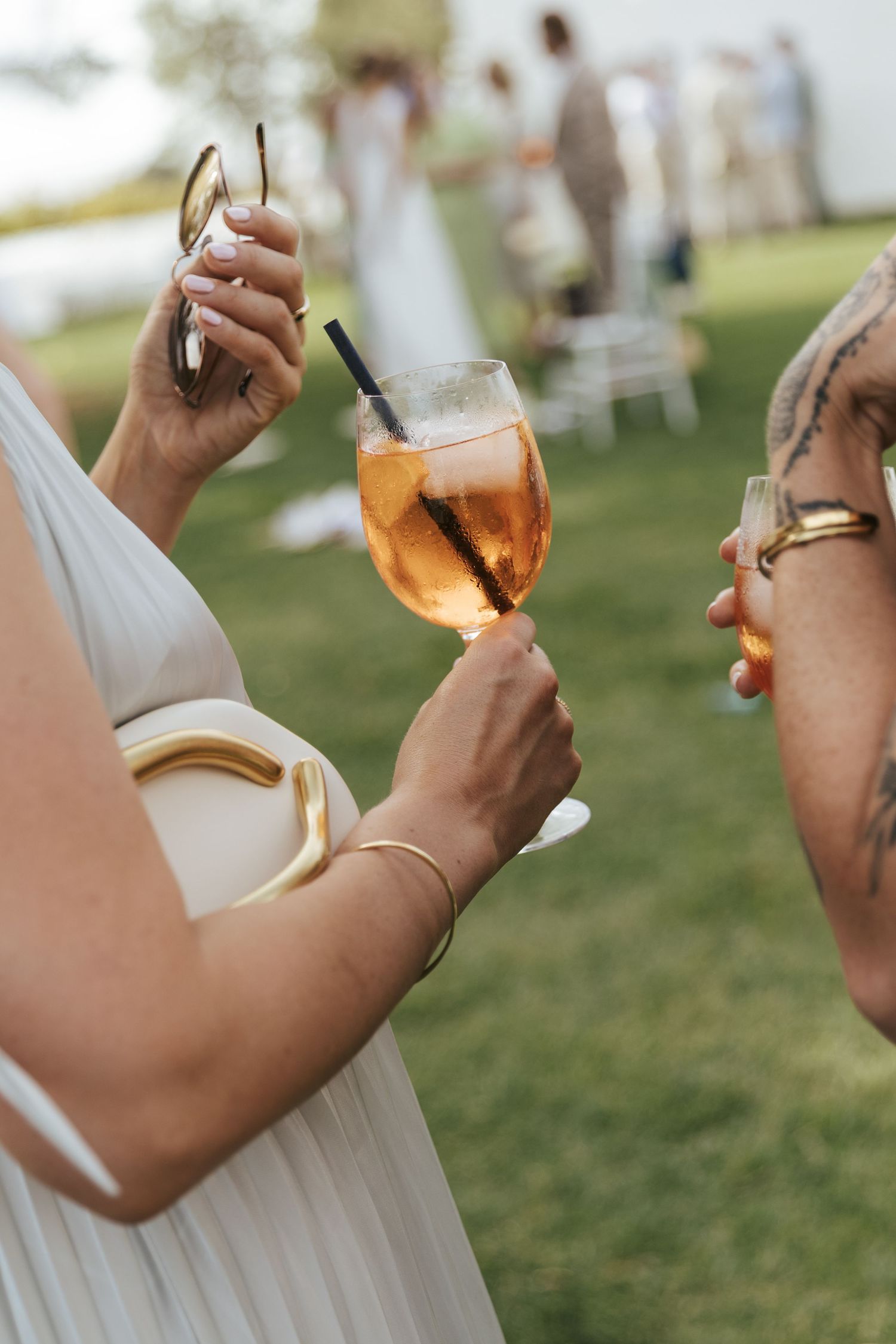 Two women holding cocktails at an outdoor wedding reception, one with a tattoo sipping a drink.