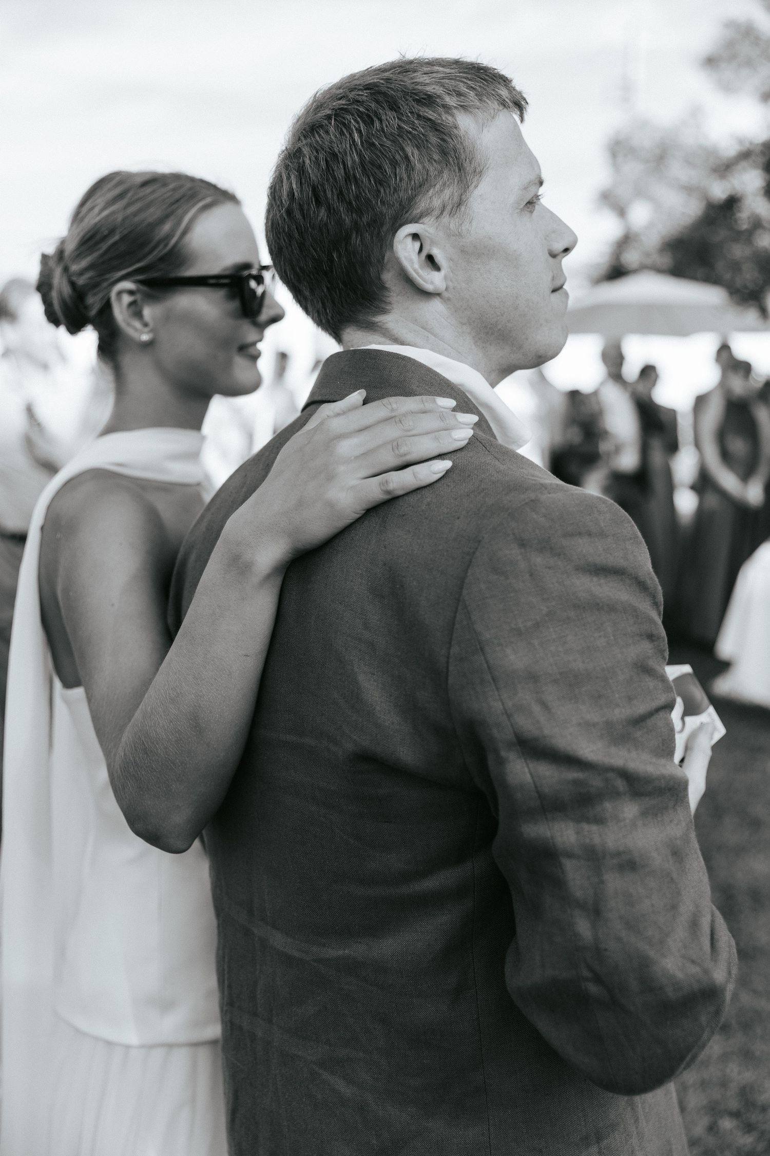 Black and white photo of a bride in sunglasses placing her hand on the groom's shoulder at a wedding.