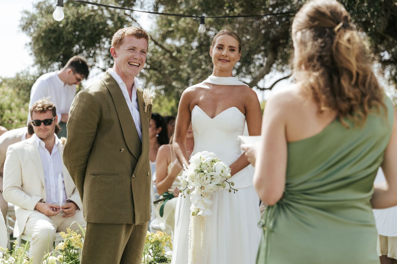 Bride in white strapless gown walks down outdoor wedding aisle with groom in tan suit while guests watch the ceremony.