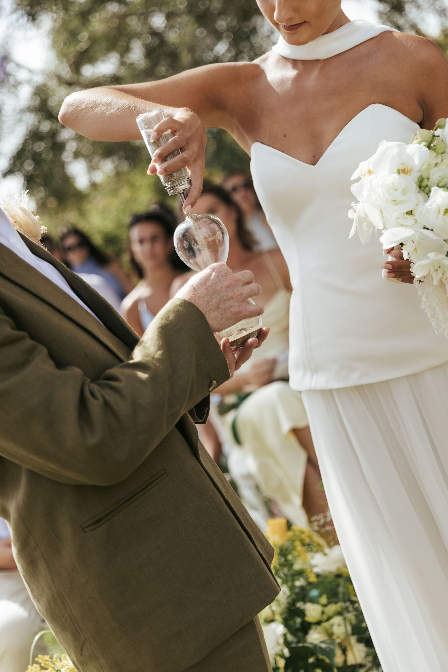 Bride in white strapless gown holding bouquet while officiant performs sand ceremony at outdoor wedding.