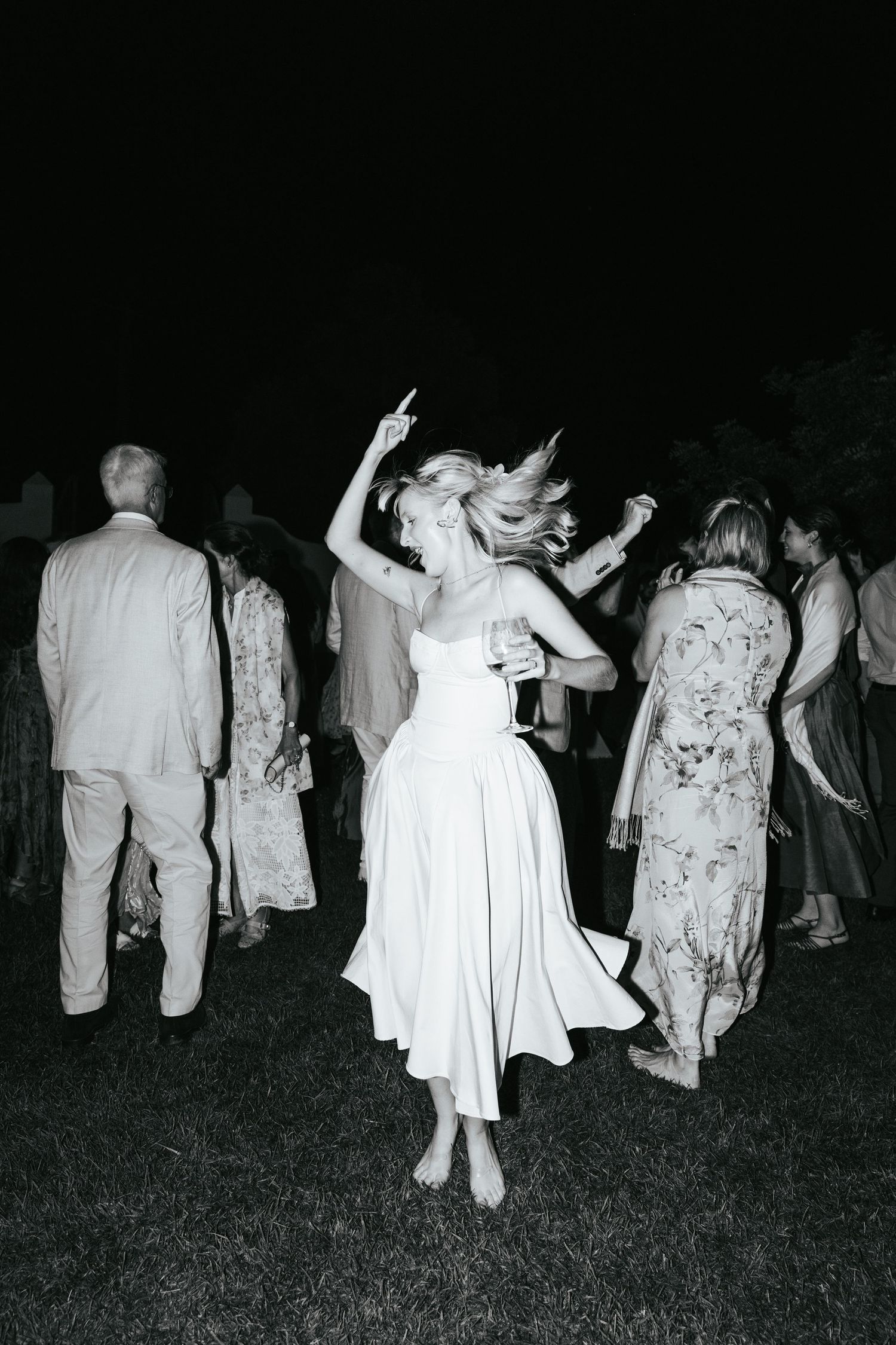 Woman in white dress dancing barefoot at an outdoor nighttime wedding reception.