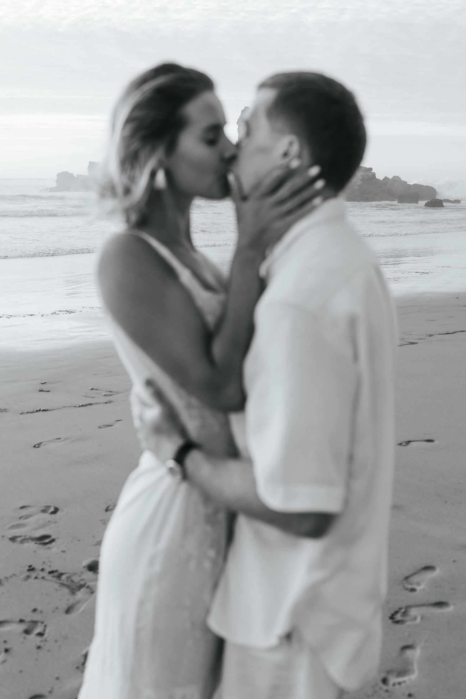 A romantic black and white photo of a couple sharing a passionate kiss on a misty beach. Praia do Castelejo, Portugal.