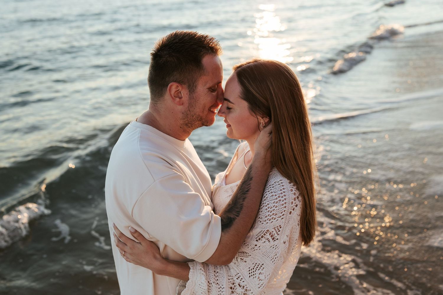 Romantic couple embraces on beach at sunset with waves and sunlight in the background.