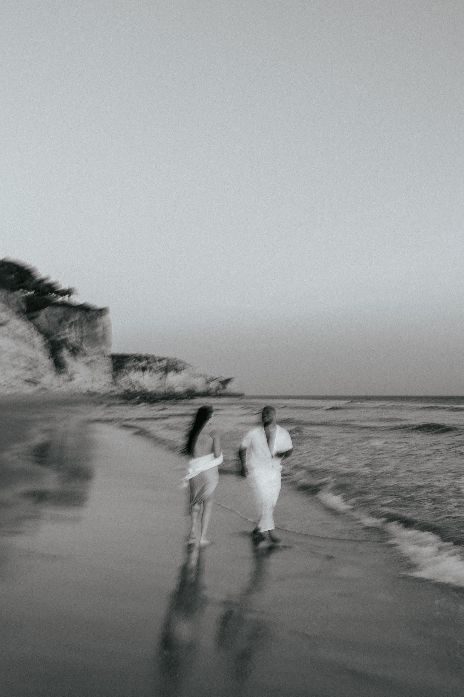 A dreamy black and white photo of two people walking along a misty beach with cliffs. Praia do Porto de Mós, Lagos, Algarve, Portugal.