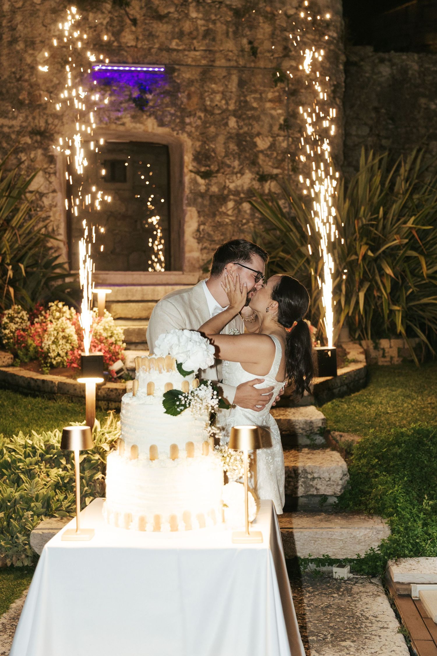 Wedding couple shares romantic moment by sparkler-lit table with white cake at stone venue.