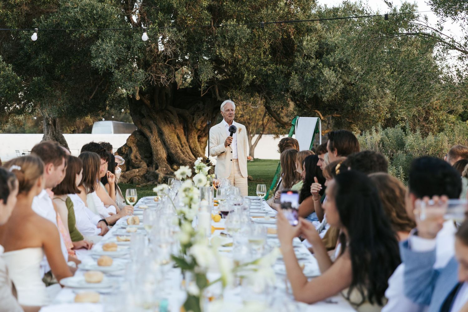 Wedding speaker addressing guests at long outdoor reception table under ancient olive tree at golden hour.