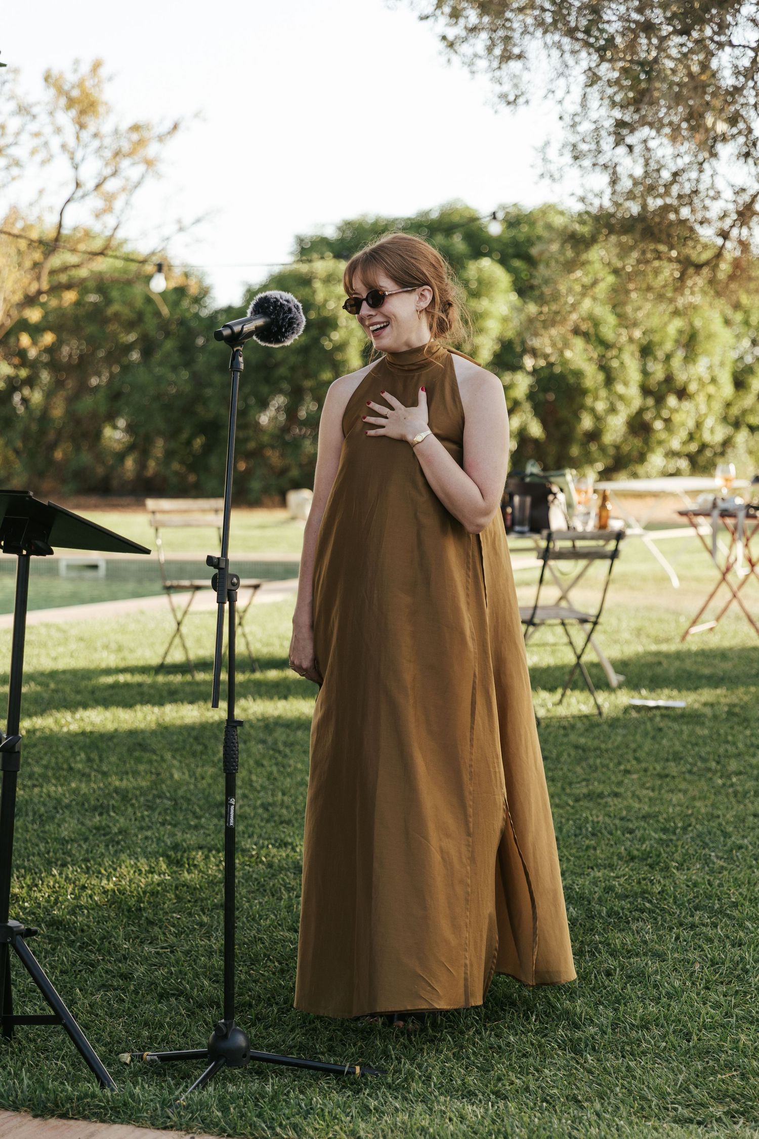 A woman in an elegant olive maxi dress stands at a microphone outdoors, smiling and touching her necklace at a garden event.