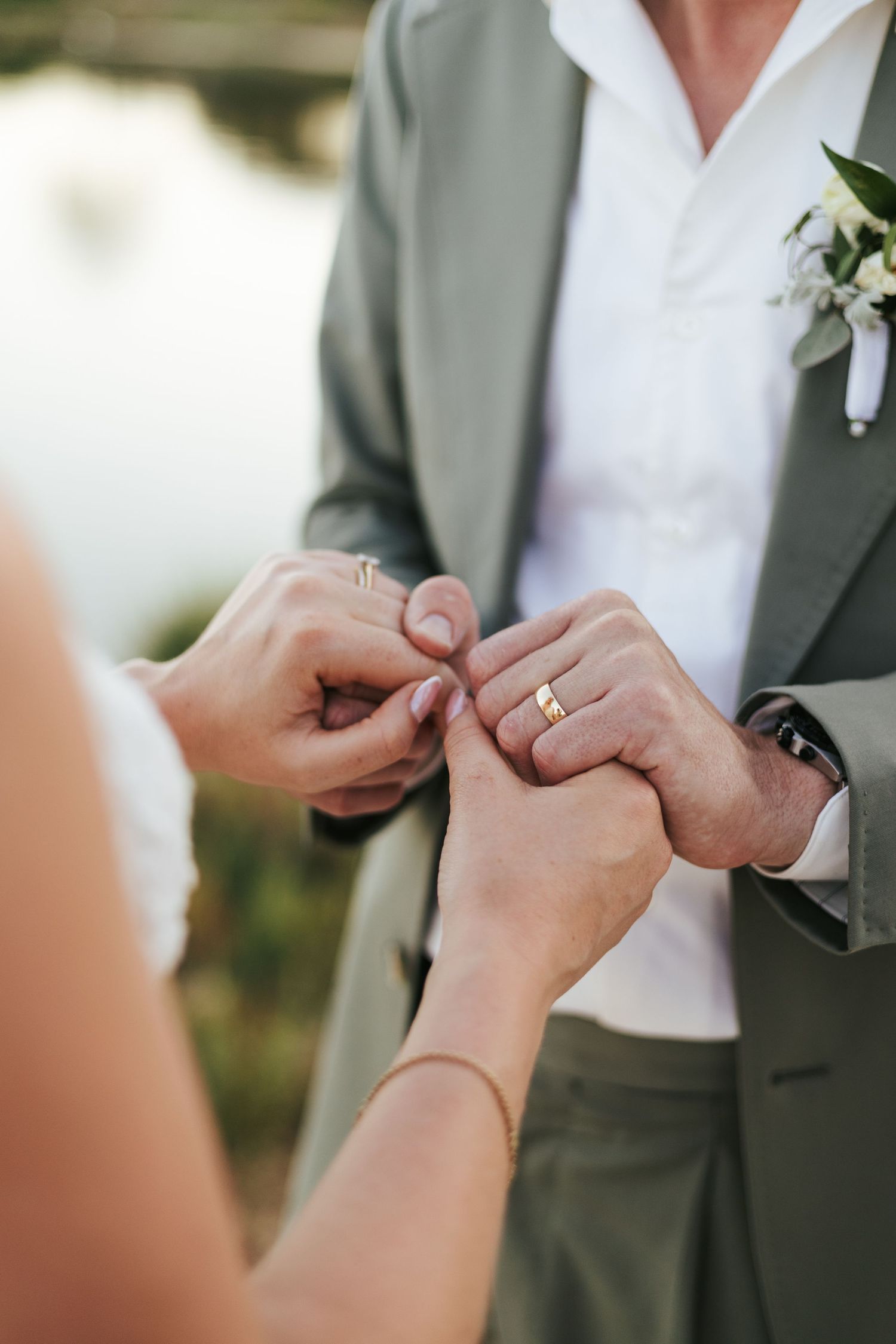 Bride placing a gold wedding ring on groom's finger during outdoor ceremony, groom wearing sage green suit with floral boutonniere.