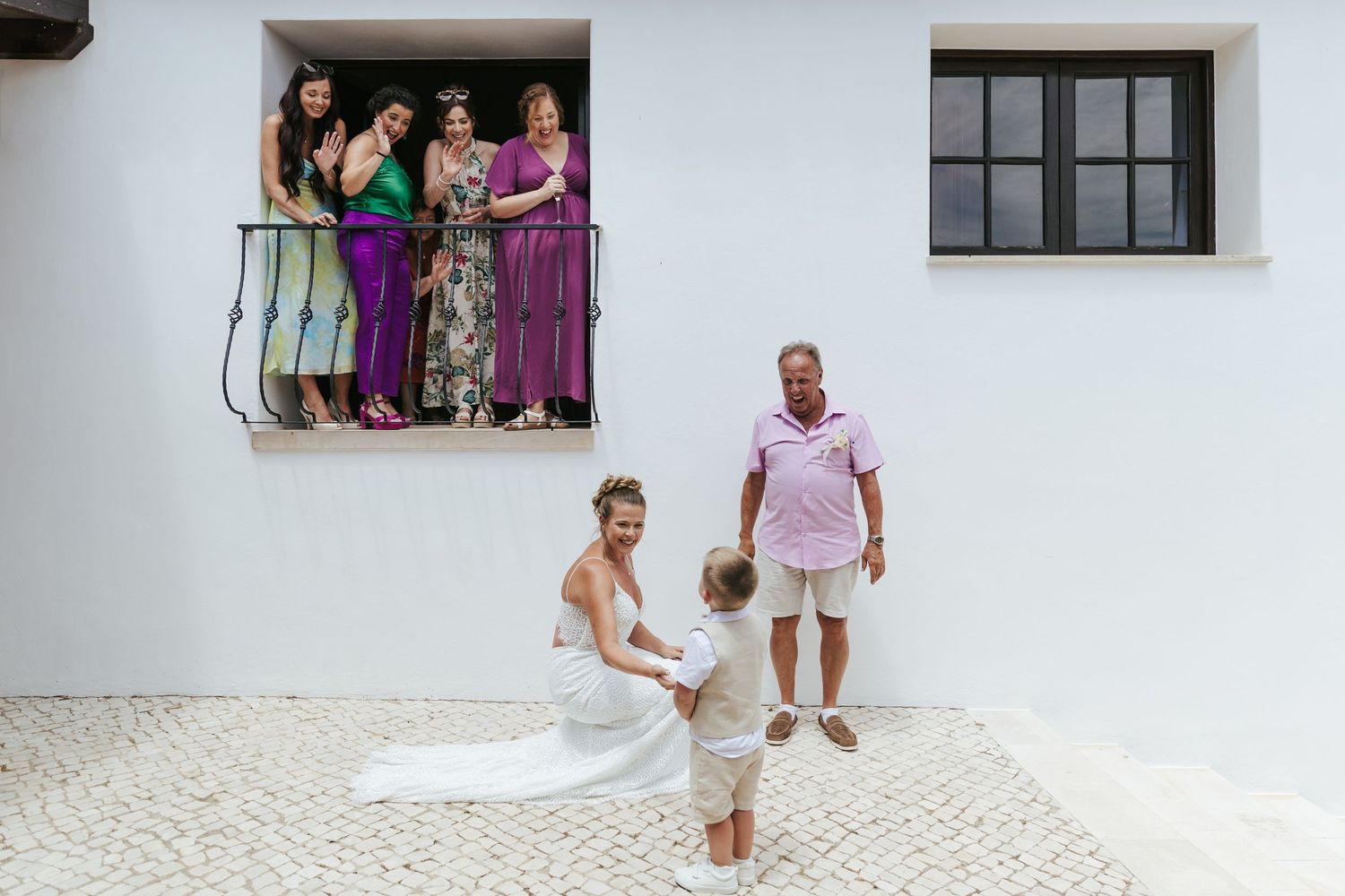 Bride in white gown crouches to greet child while bridesmaids in purple watch from balcony at outdoor wedding venue.