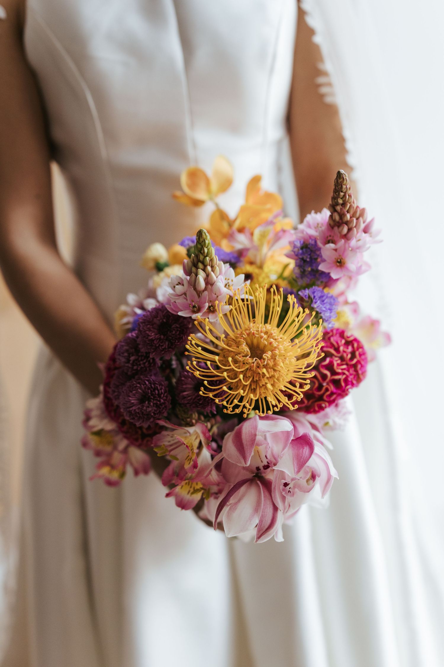 Bride in white gown holding a vibrant wedding bouquet with yellow, pink, and purple flowers including protea and hyacinth.