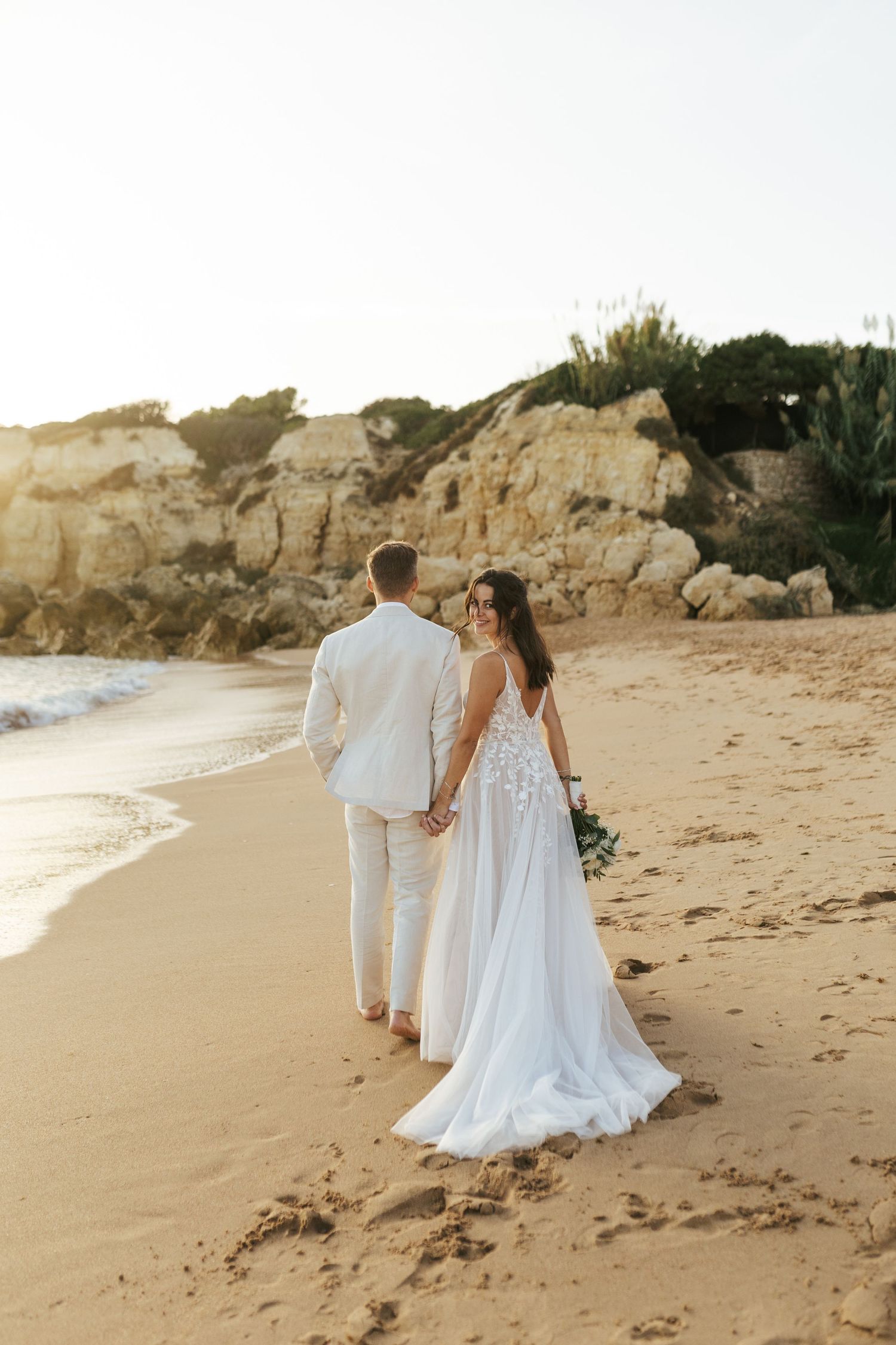 Newlywed couple walking on sandy beach at sunset in white wedding attire.