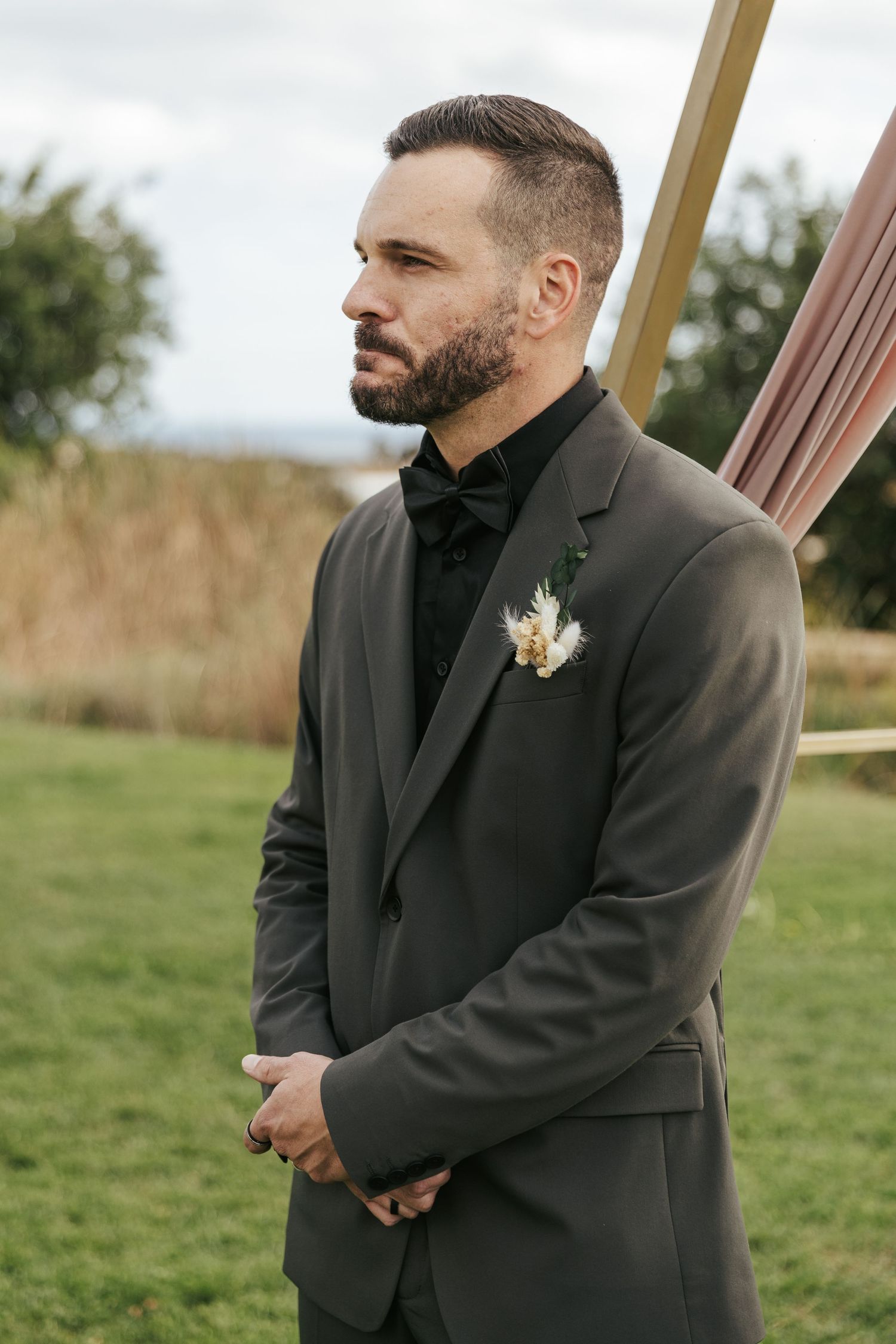 A groom wearing a dark suit and boutonniere stands thoughtfully during an outdoor wedding ceremony.