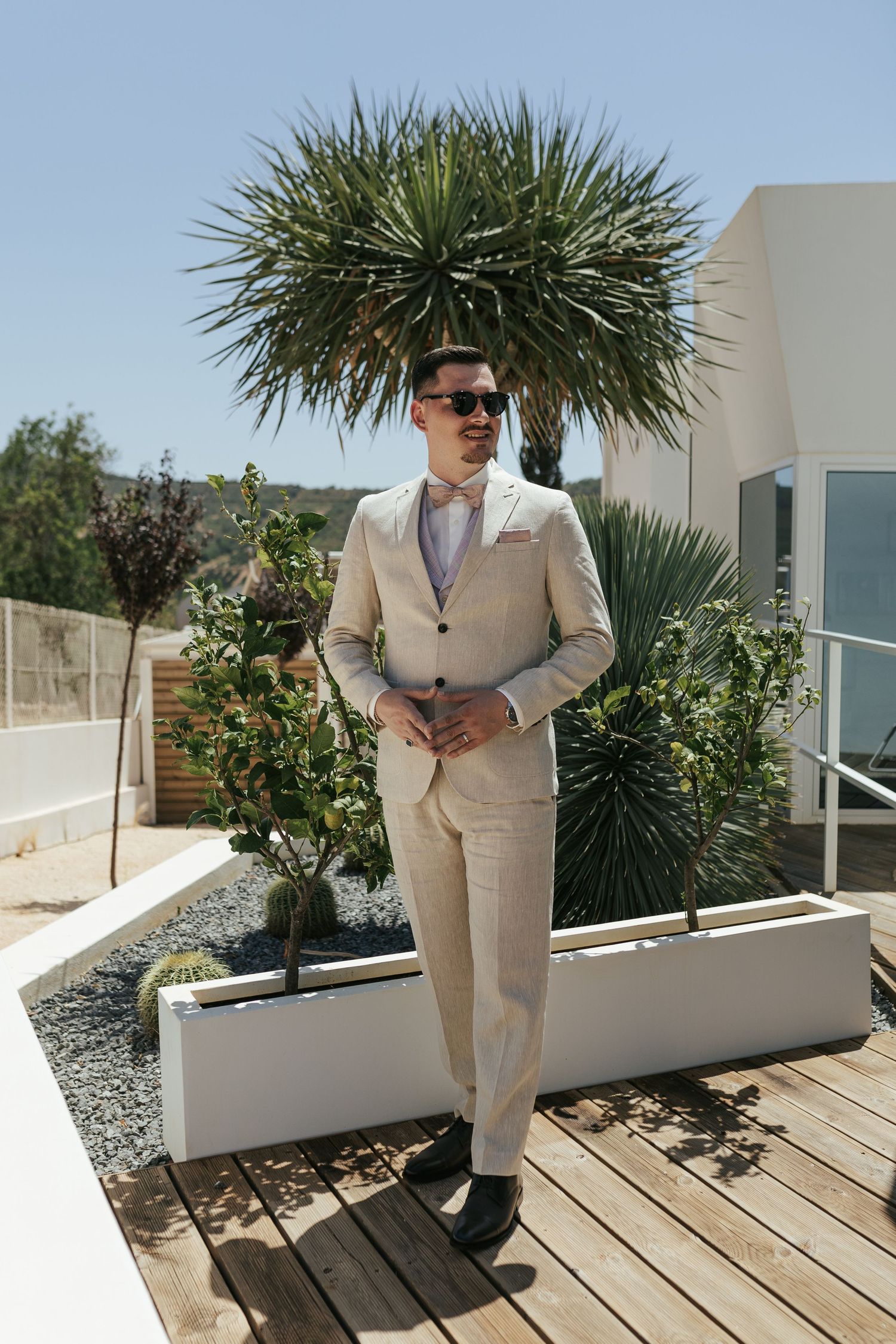 Elegant beige summer suit worn at tropical outdoor venue with palm trees and wooden deck in background.