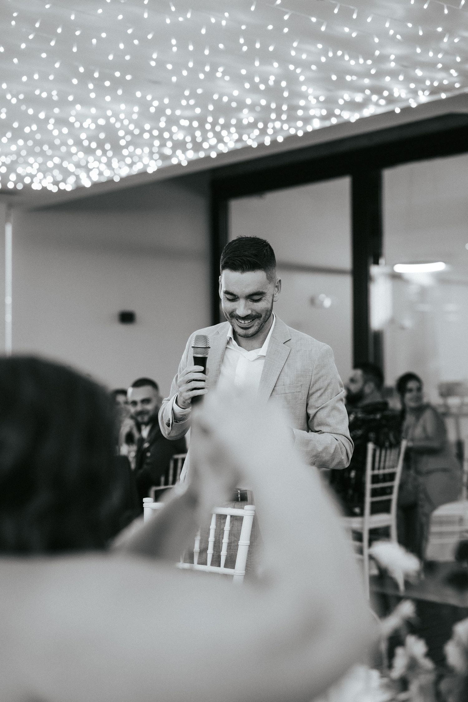 A speaker gives a speech under twinkling string lights at an indoor reception venue.