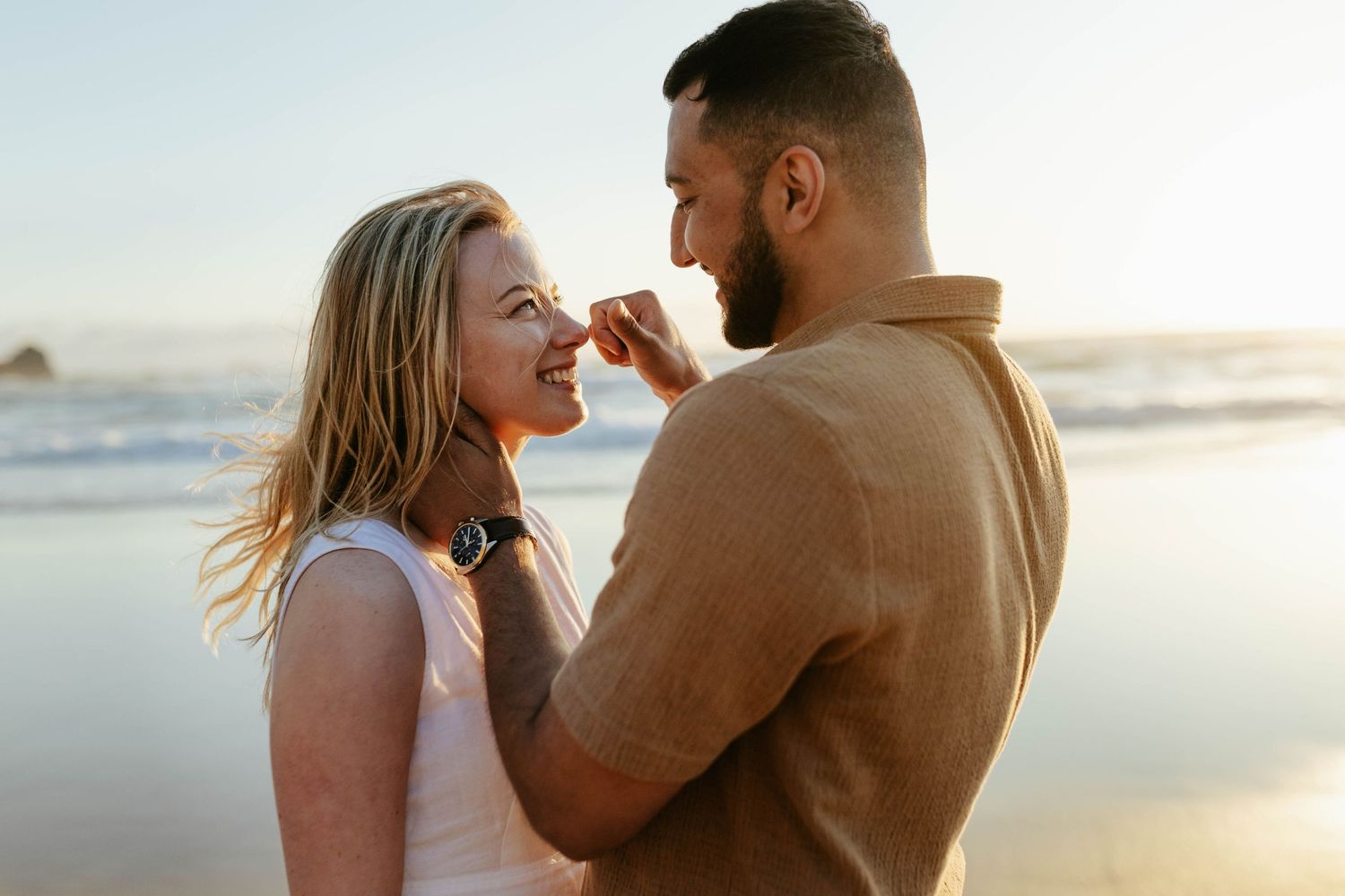 A joyful couple shares a playful moment while standing on a beach with golden sunlight and ocean waves behind them.