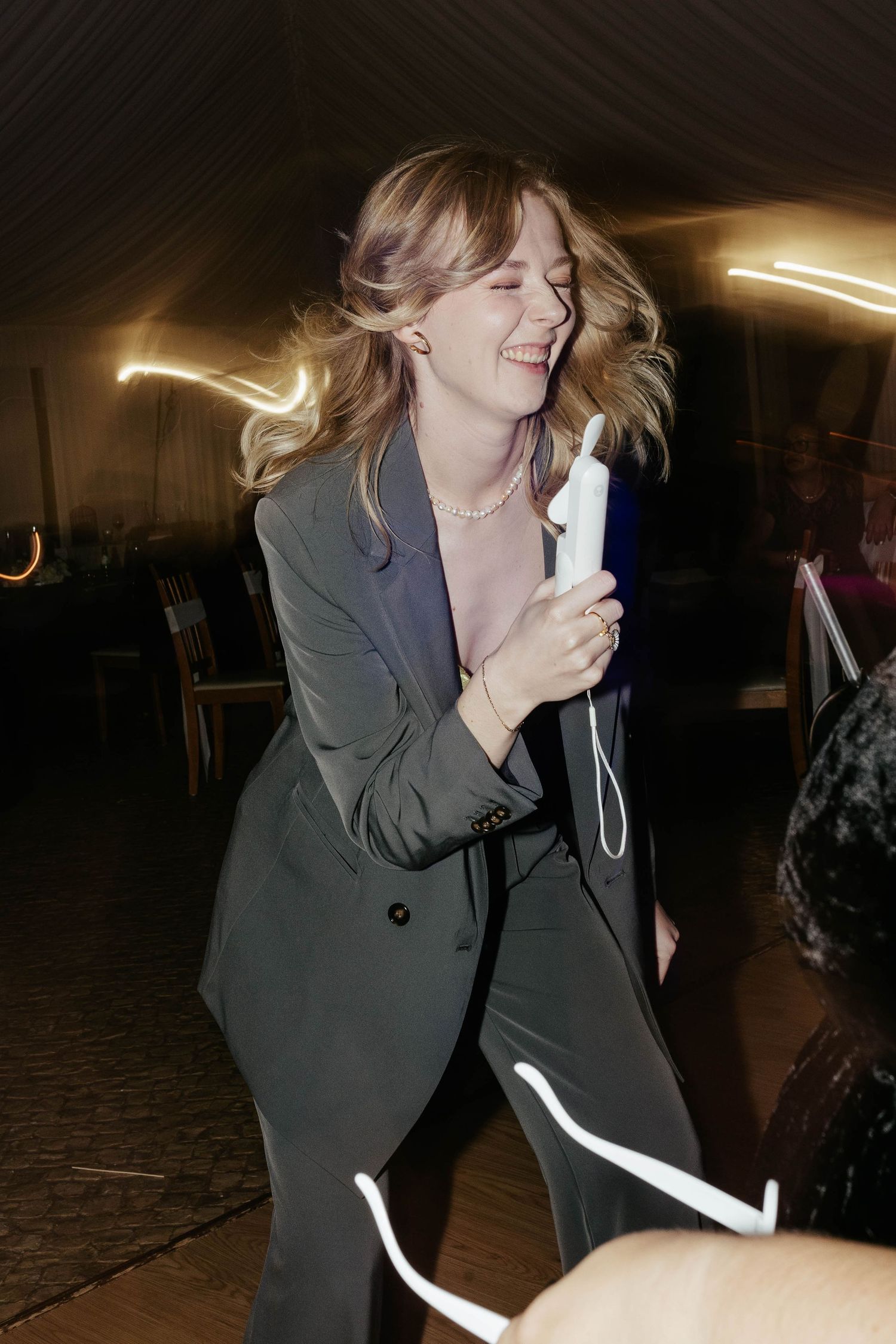 A person in a dark blazer laughs joyfully while holding a microphone in a dimly lit venue.