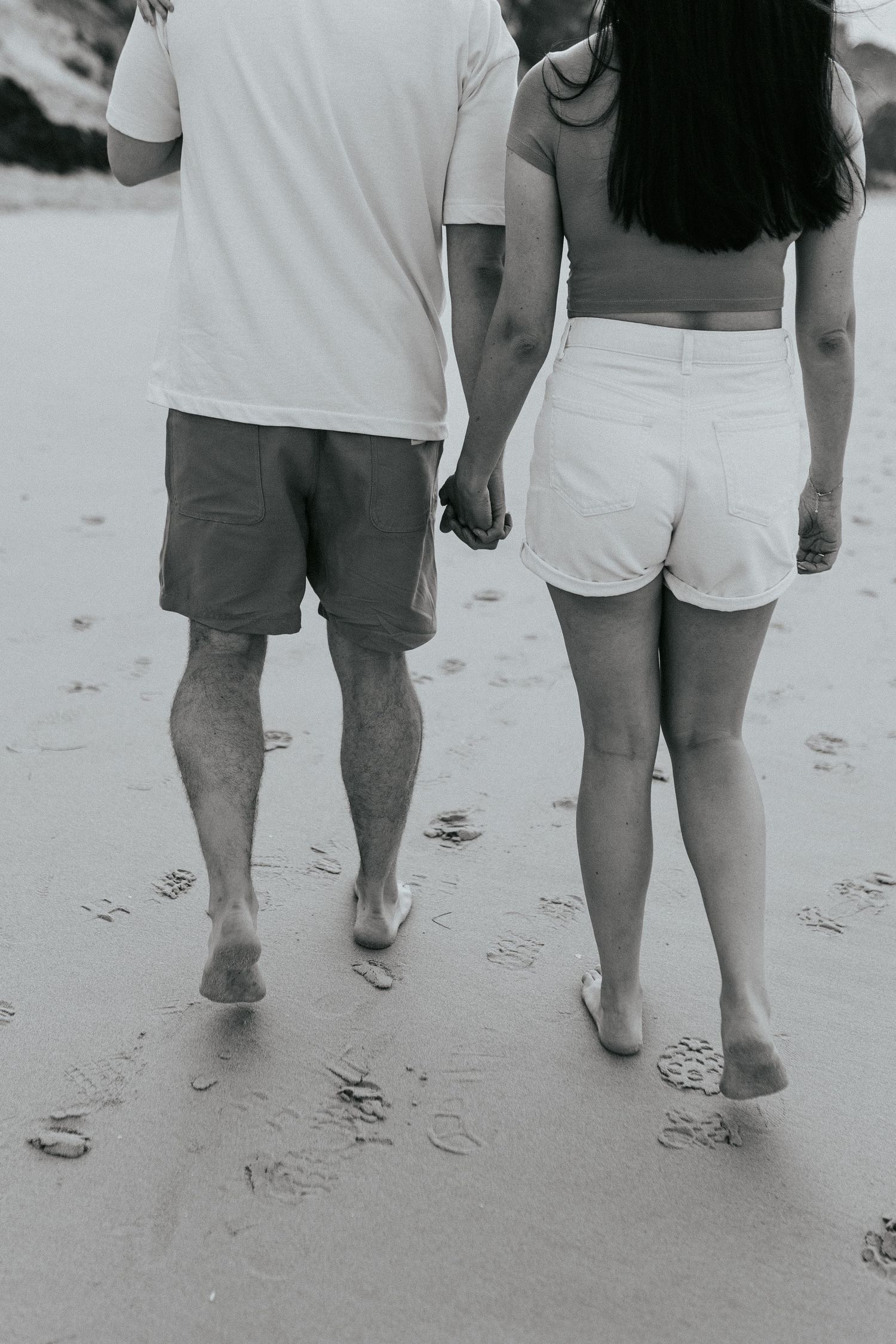 A couple walking barefoot on a sandy beach holding hands, wearing white shorts and t-shirt, viewed from behind in black and white.