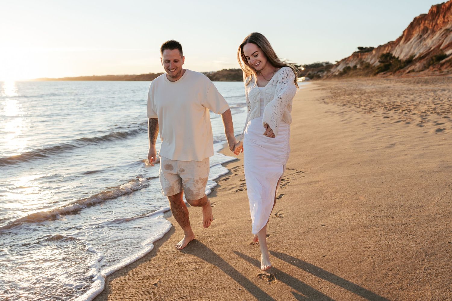 Couple walking hand in hand along a sunset beach in casual white outfits.