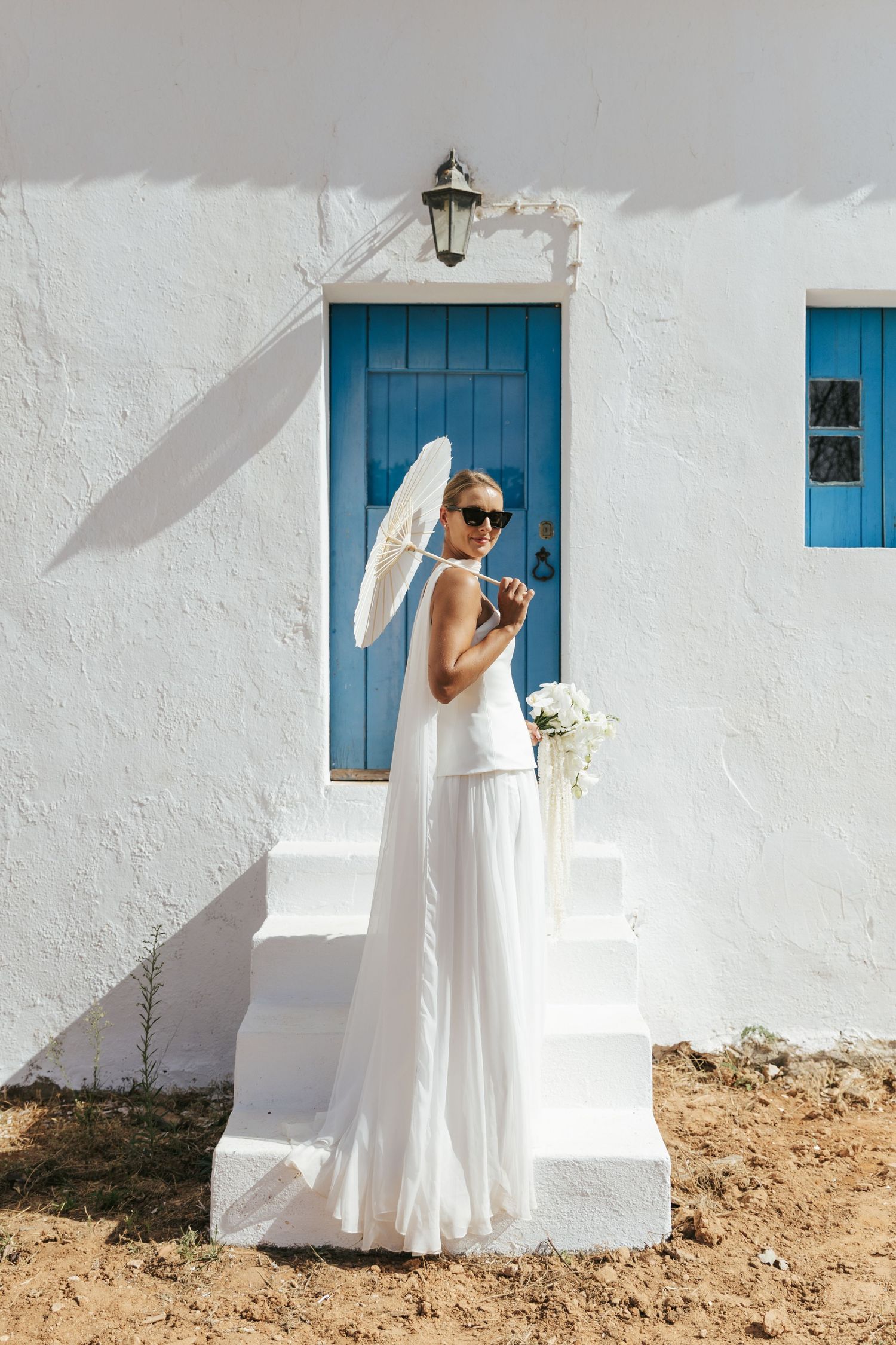 Bride in white dress holding parasol and bouquet stands on steps before blue door of whitewashed building.