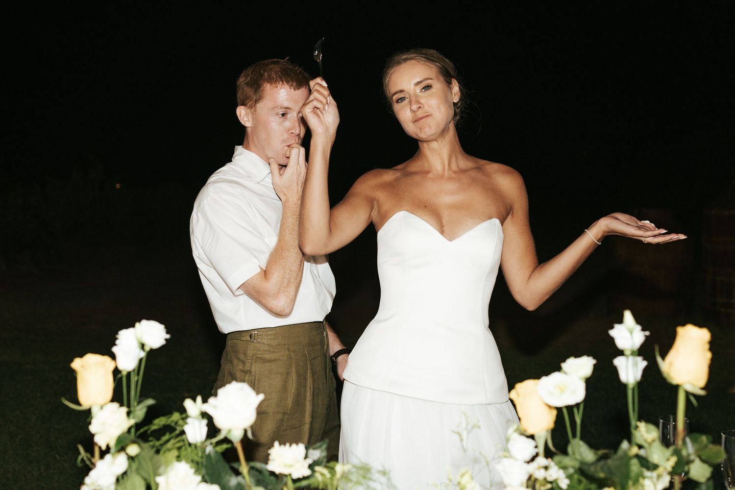 Bride and groom posing playfully at night surrounded by white and yellow roses at their wedding celebration.