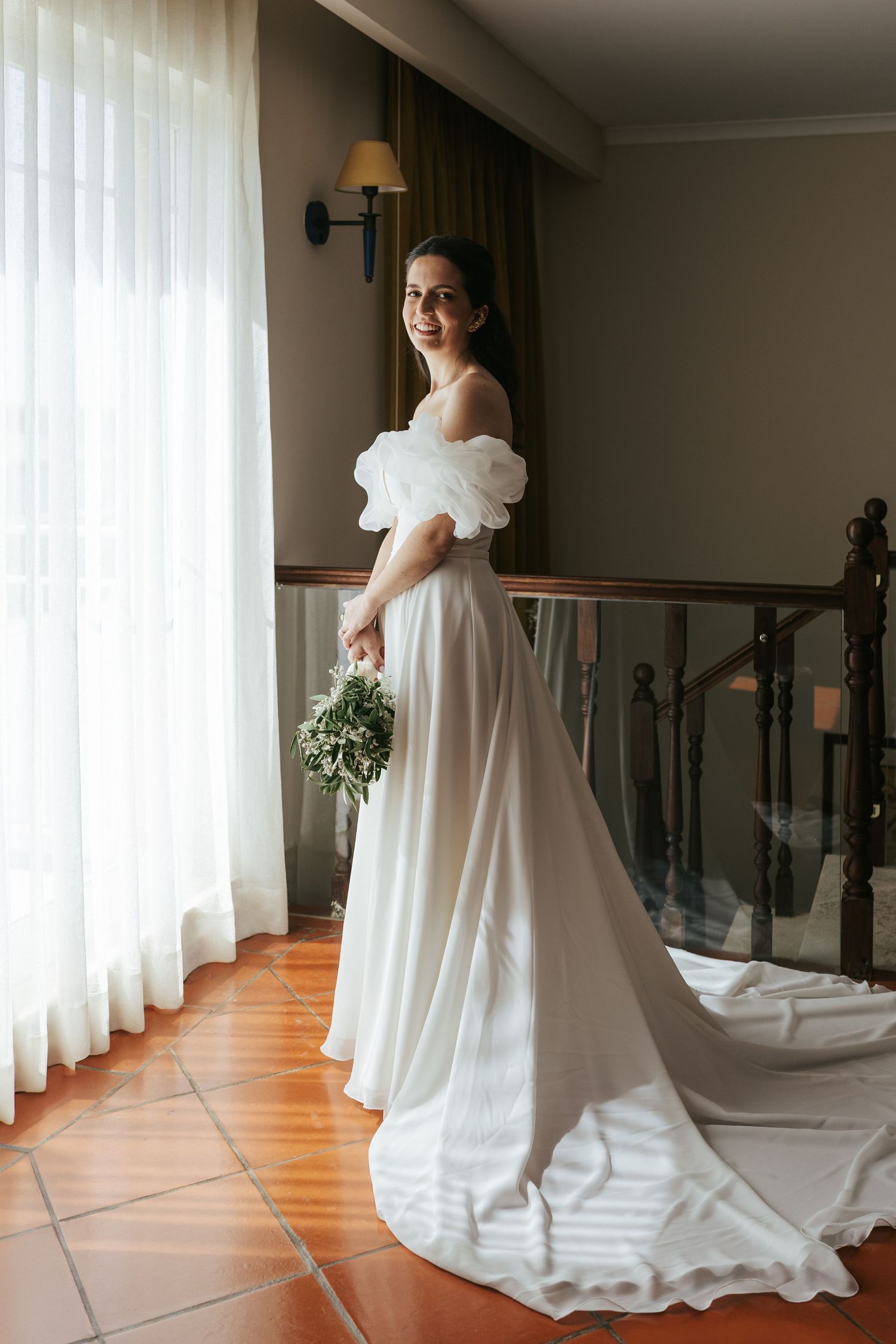 A white wedding gown with dramatic ruffled sleeves and flowing train displayed in a sunlit room with hardwood floors.