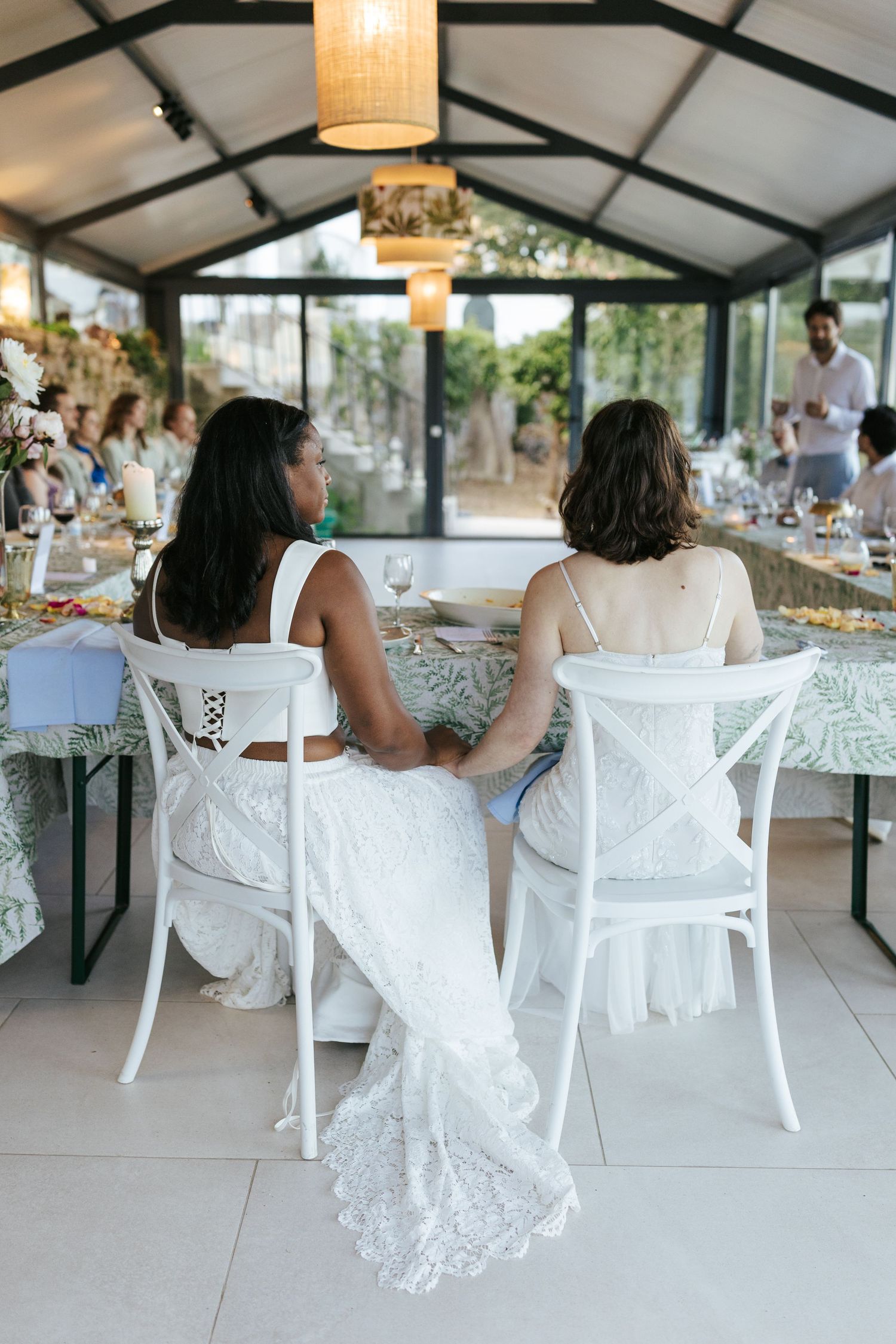 Two brides in white dresses sit together at a wedding reception table in an elegant glass-roofed venue.