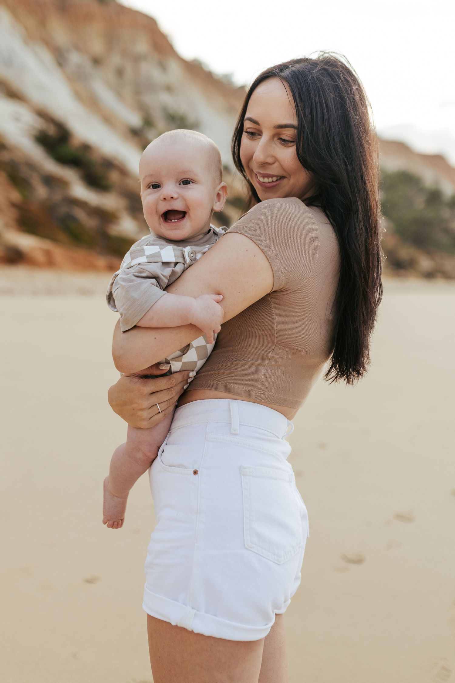 Parent in brown top and white shorts holds laughing baby on sunny beach with orange cliffs behind them.