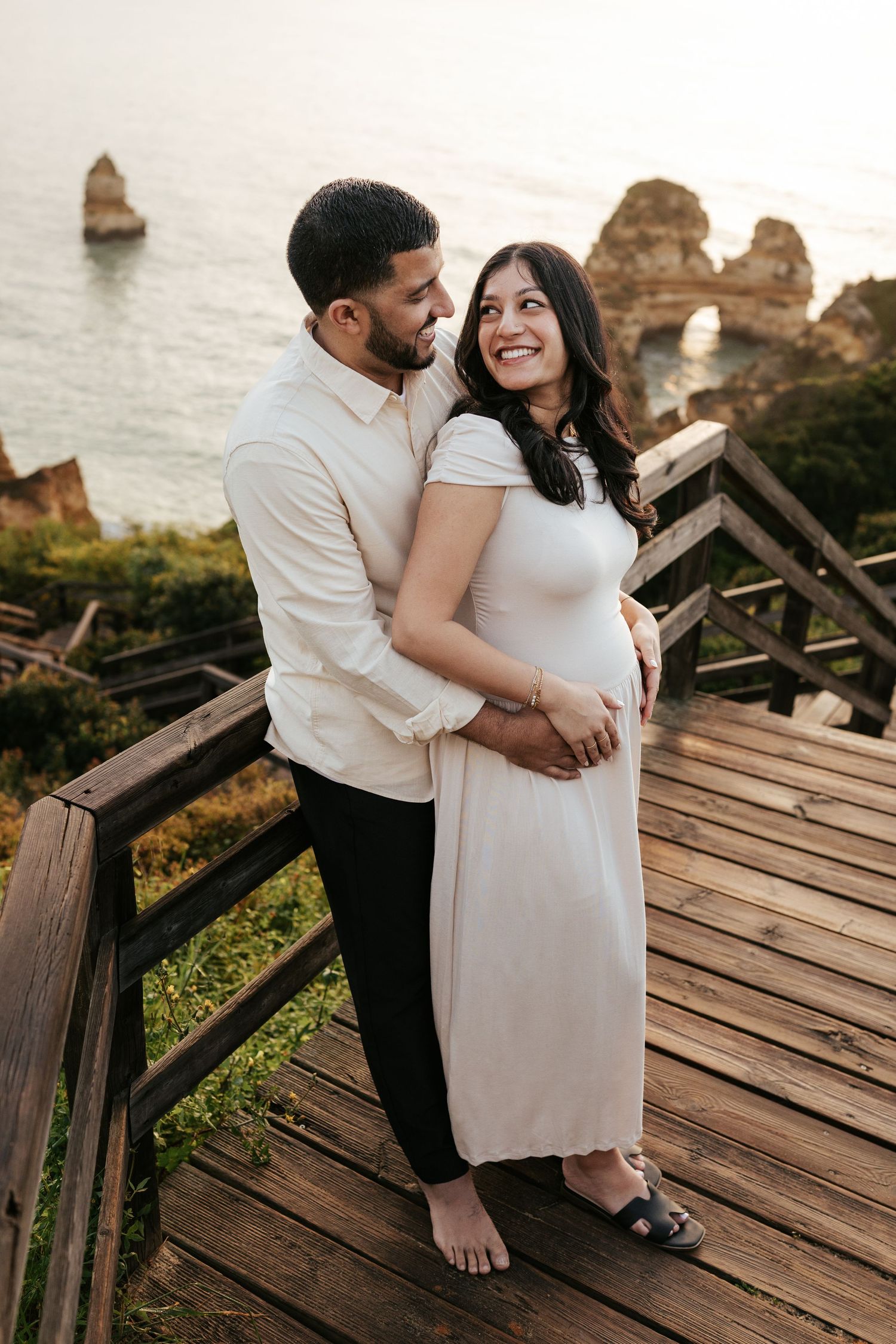 Expecting couple embraces on wooden deck overlooking coastal rock formations.