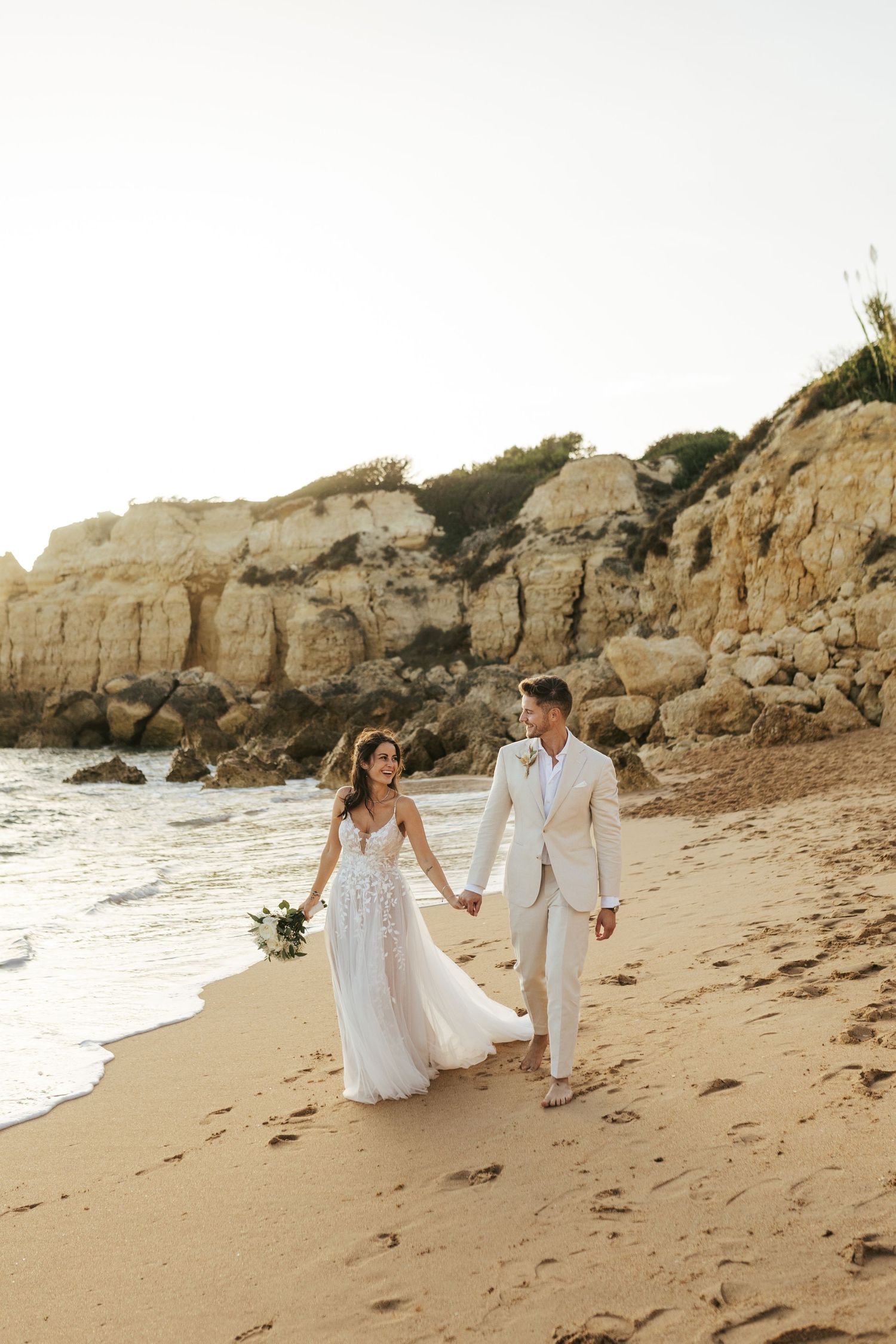 A couple walks hand in hand on a sandy beach with dramatic cliffs and ocean waves at sunset.