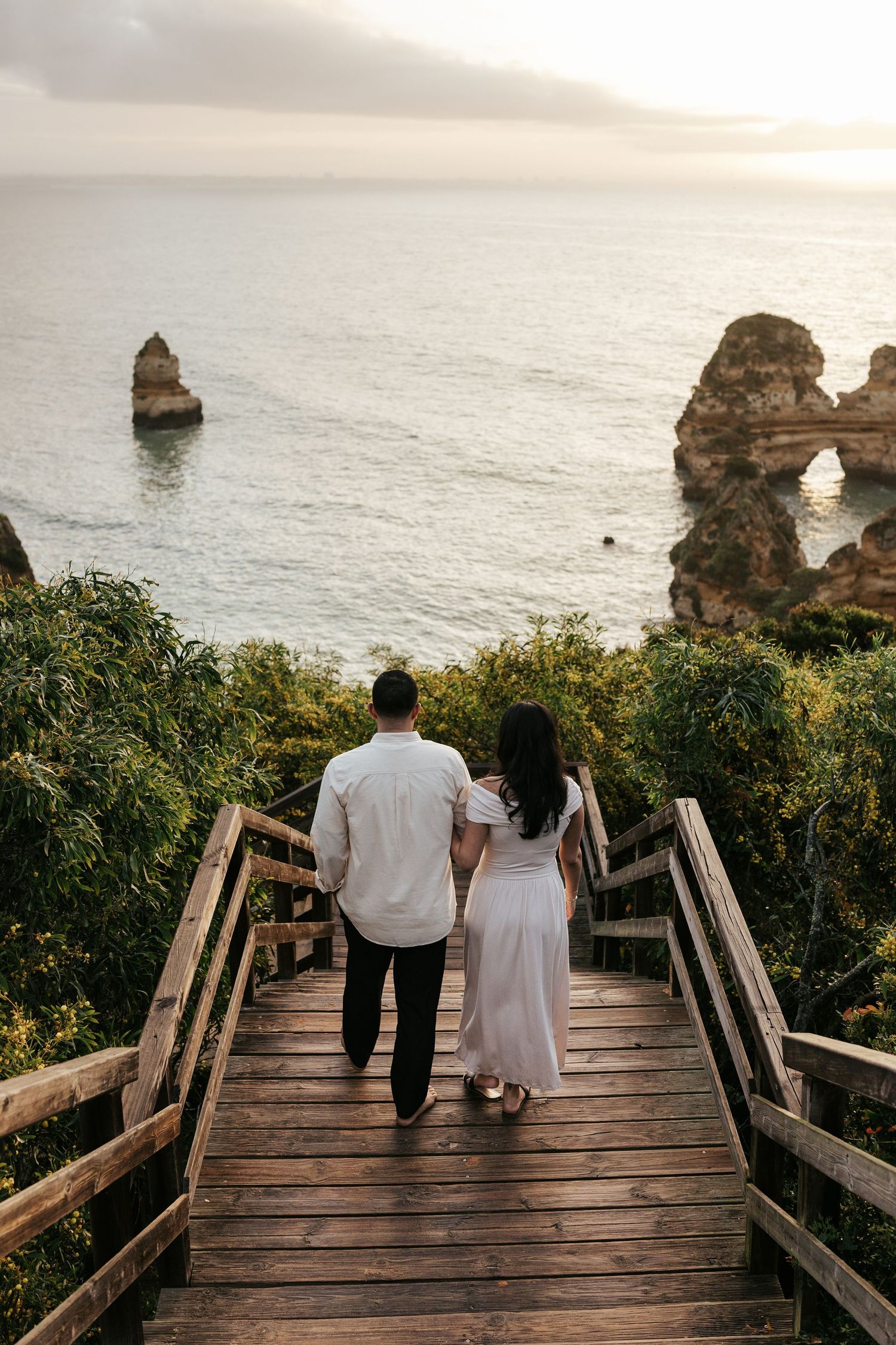 Couple walking down wooden boardwalk stairs overlooking dramatic coastal rock formations at sunrise. Praia do Camilo, Lagos, Algarve, Portug