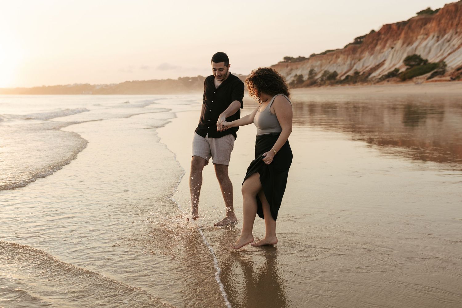 Two people standing together at the beach during sunset with cliffs in the background.