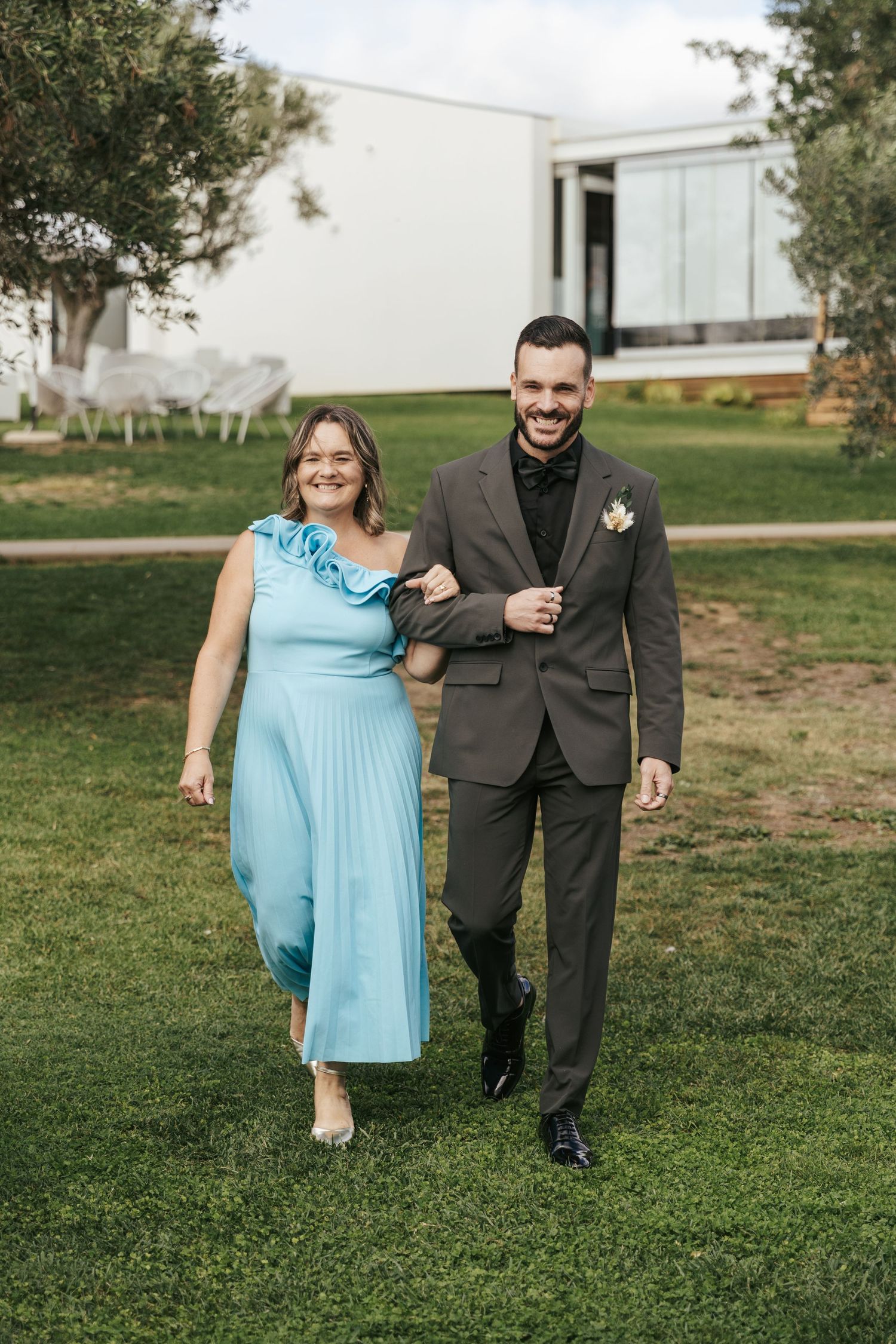 A couple walks together on grassy grounds outside a modern white building, with the person wearing powder blue and formal black attire.