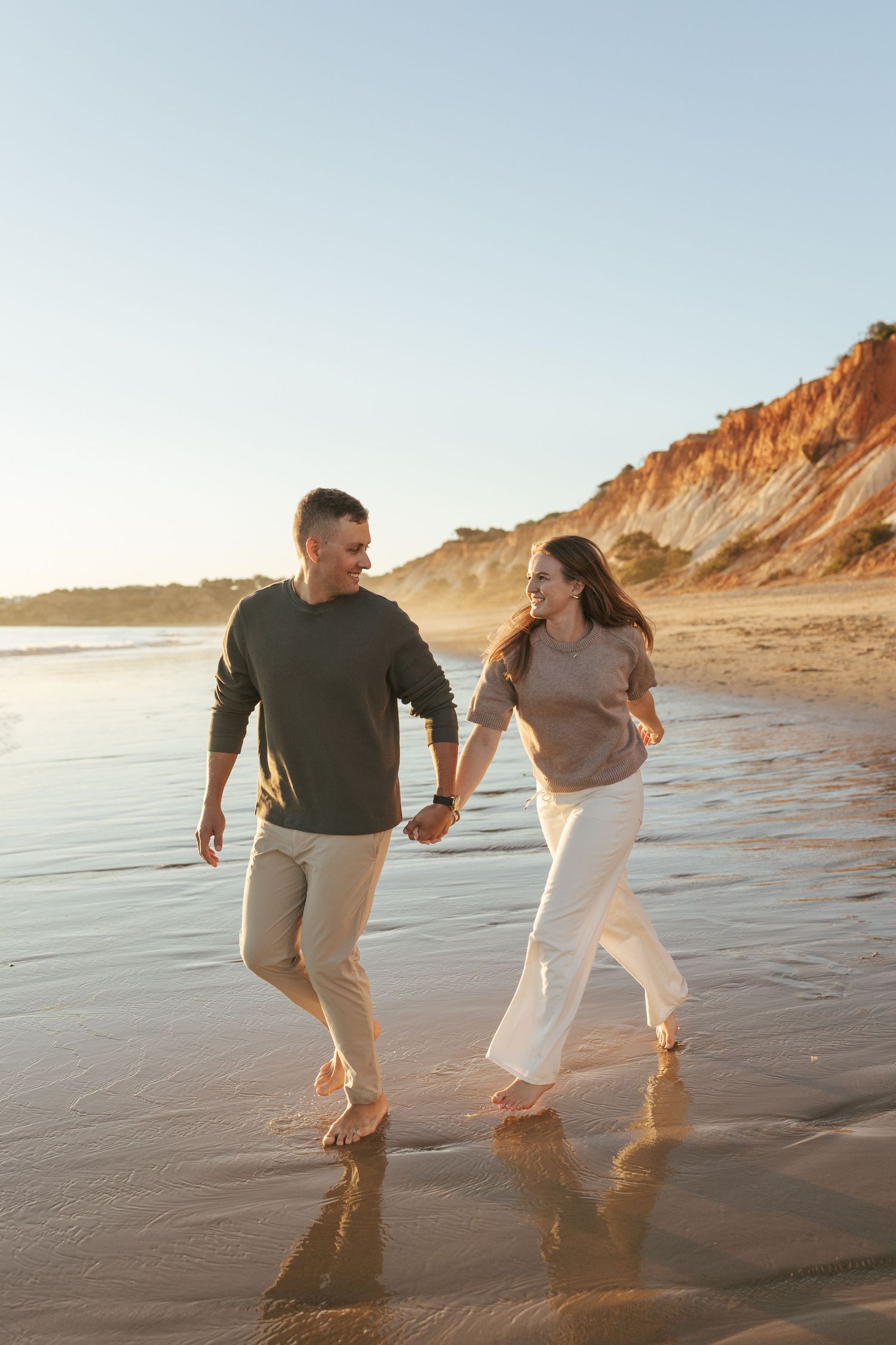 A couple walking and laughing together on a sandy beach at sunset with cliffs in the background.