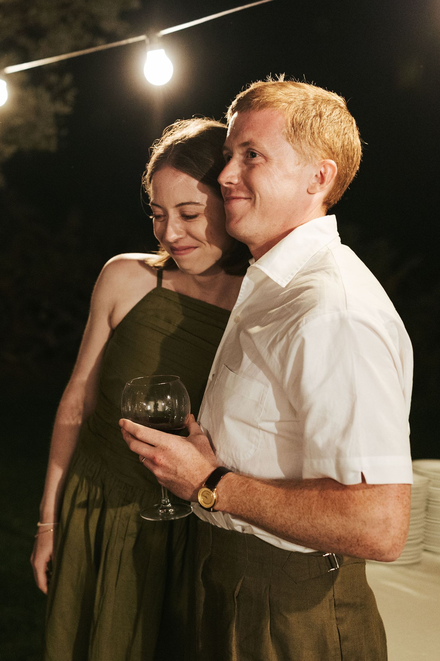 Romantic couple embracing at outdoor evening wedding reception holding red wine glass.