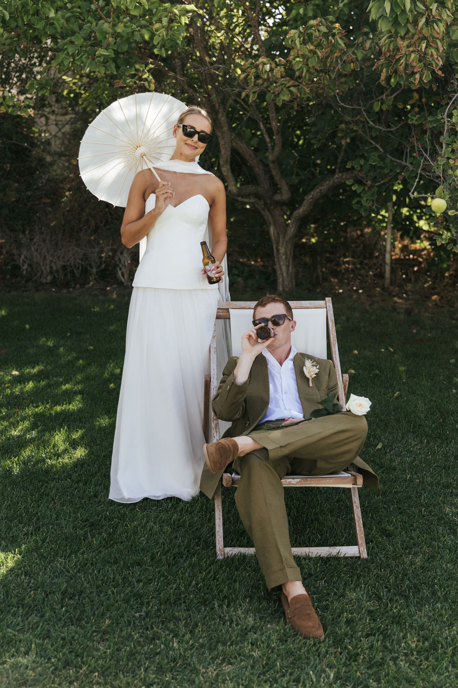 Bride in white gown holds parasol while groom sits in deck chair with camera at outdoor garden wedding.
