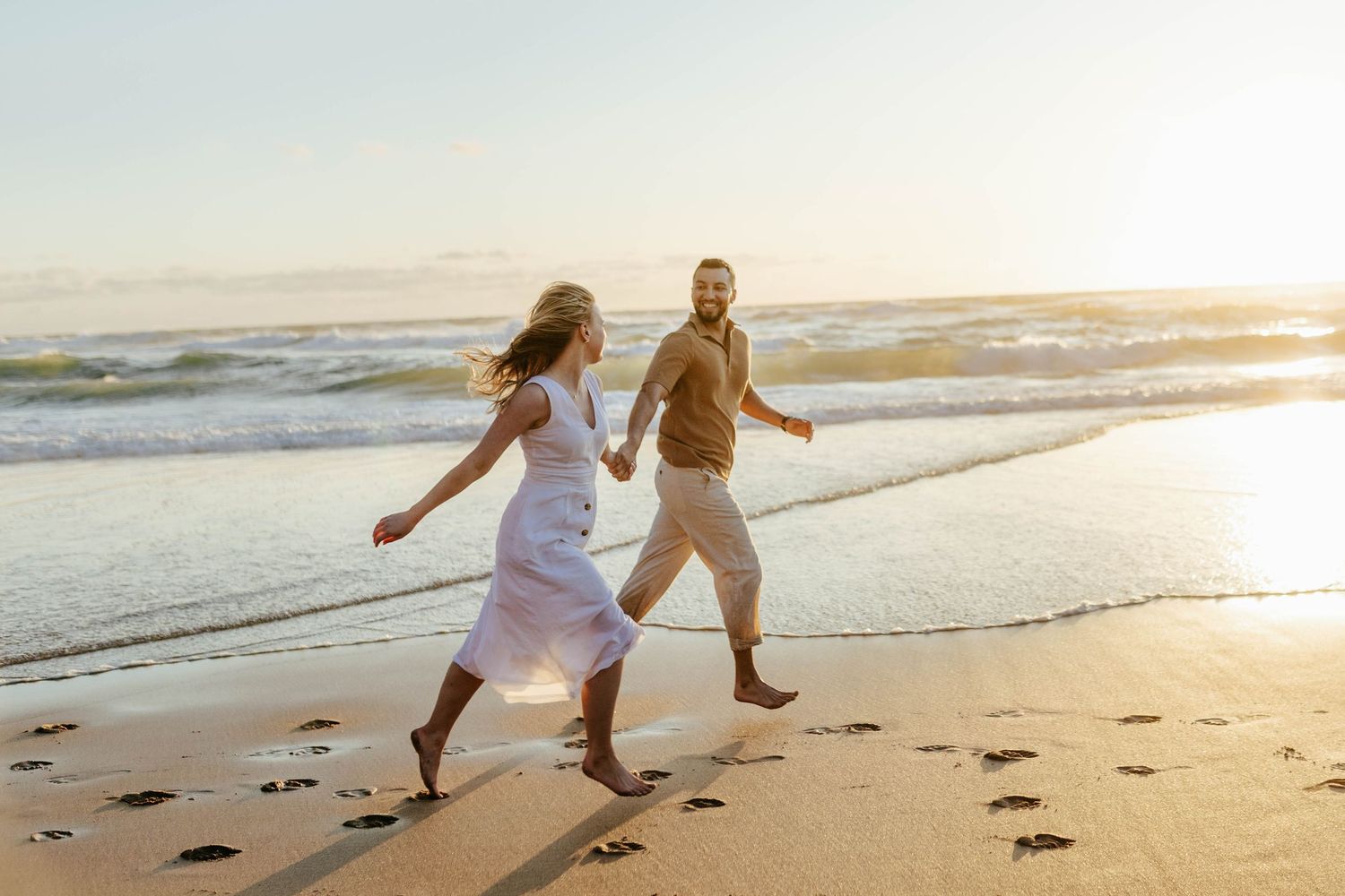 Two people running playfully along the beach at sunset, leaving footprints in the sand behind them.