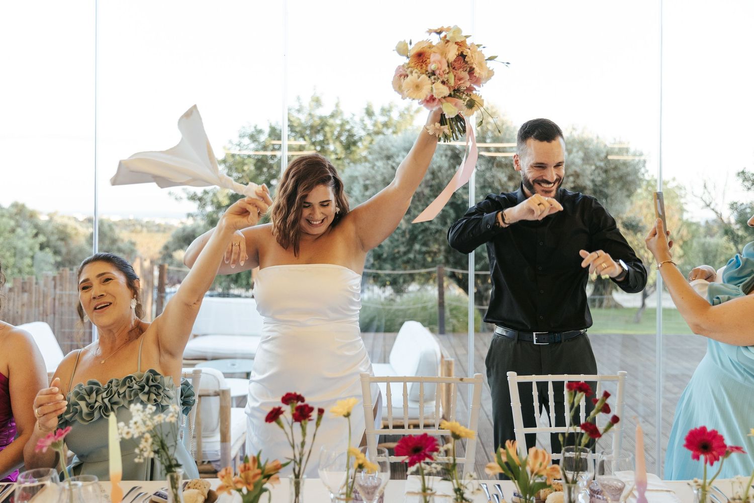 Wedding party celebrates joyfully at an outdoor reception with raised arms and floral centerpieces on the table.