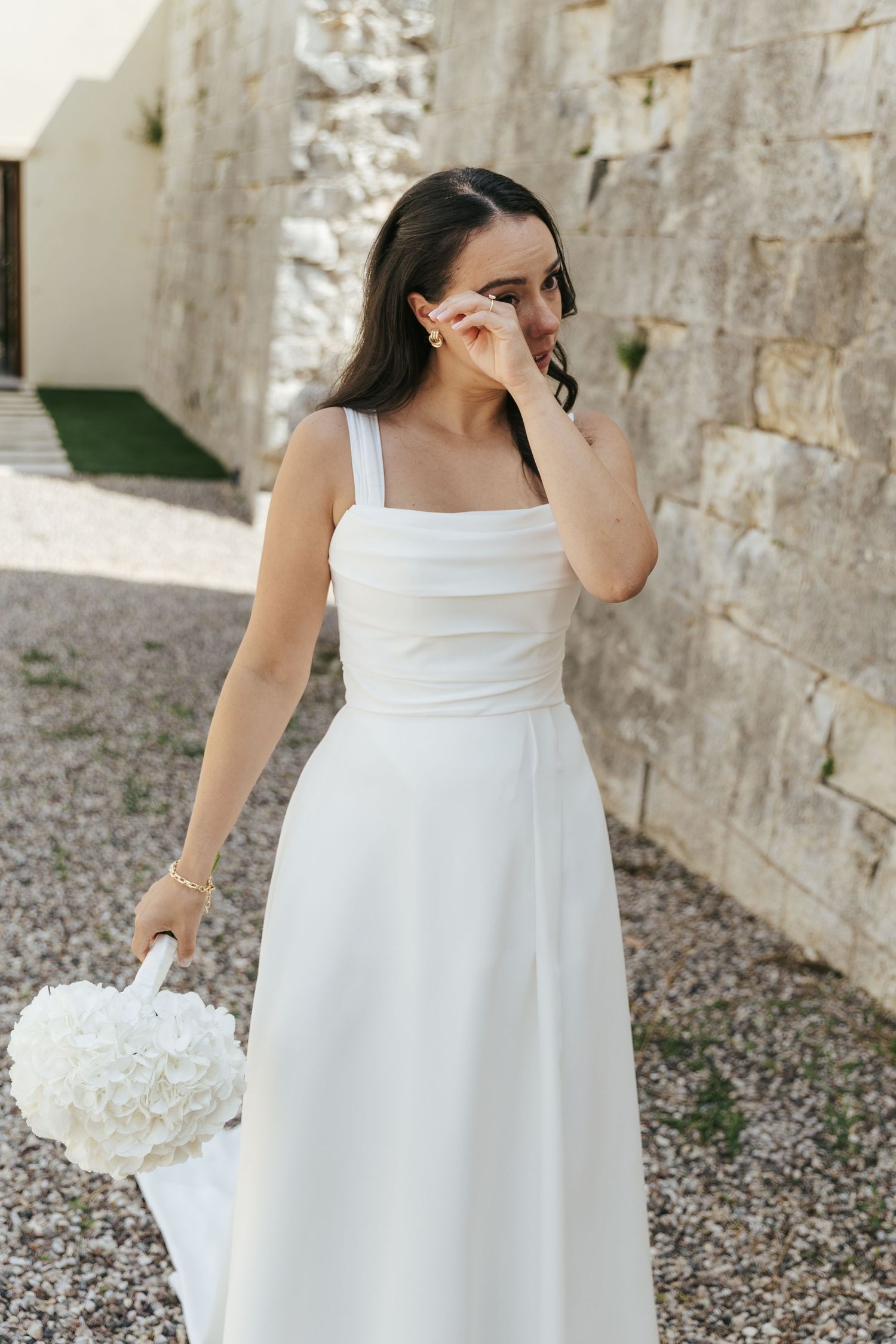 A bride in a classic white gown holds a bouquet while posing against a stone wall.
