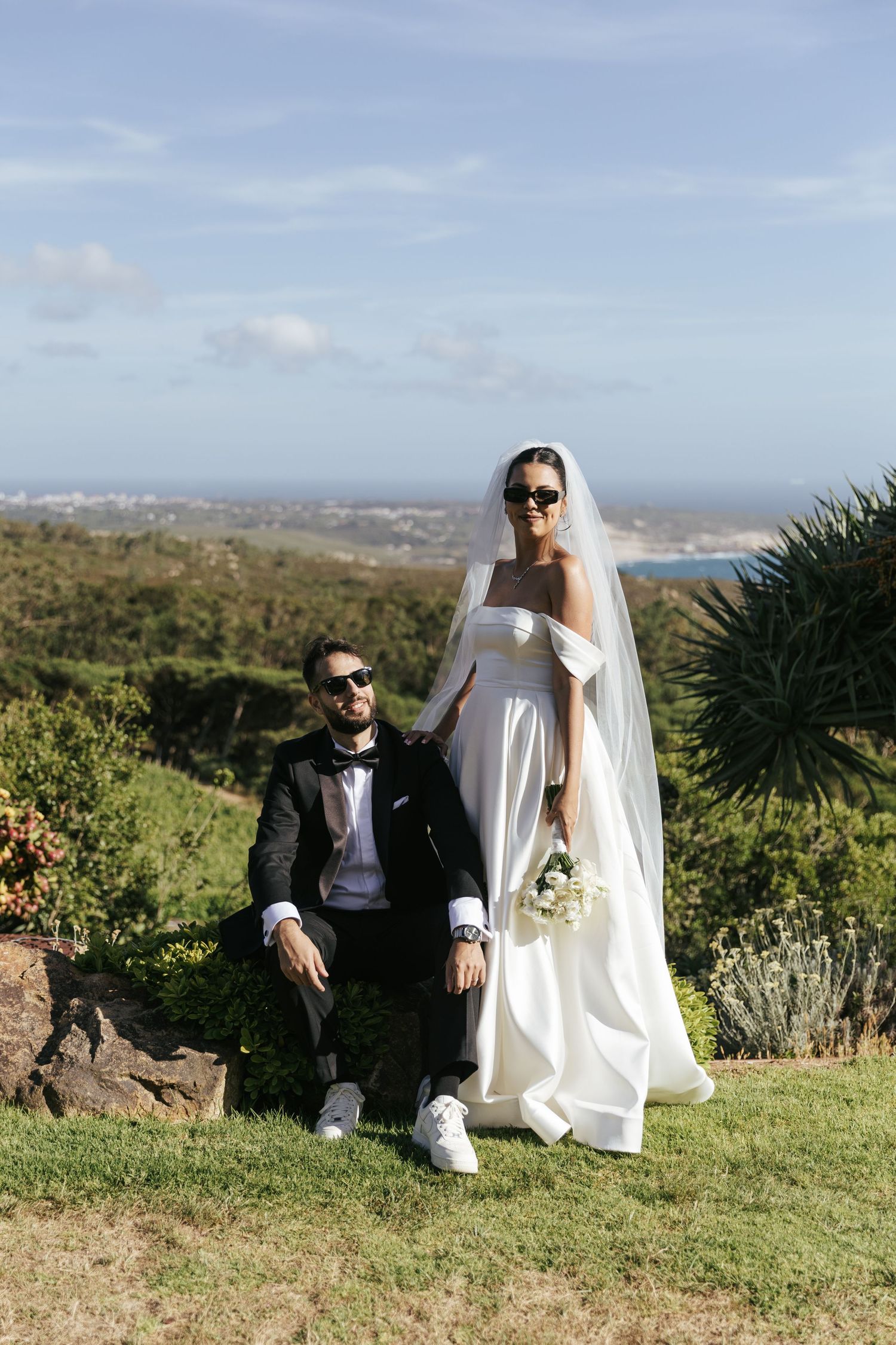 Bride in white gown and veil with groom in black tuxedo posing outdoors with ocean views on their wedding day.