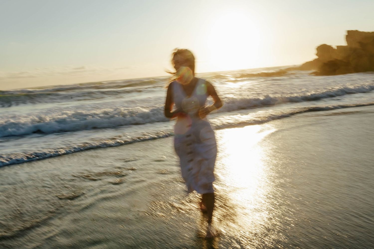 A silhouetted figure runs along the water's edge on a beach at sunset with waves crashing nearby.