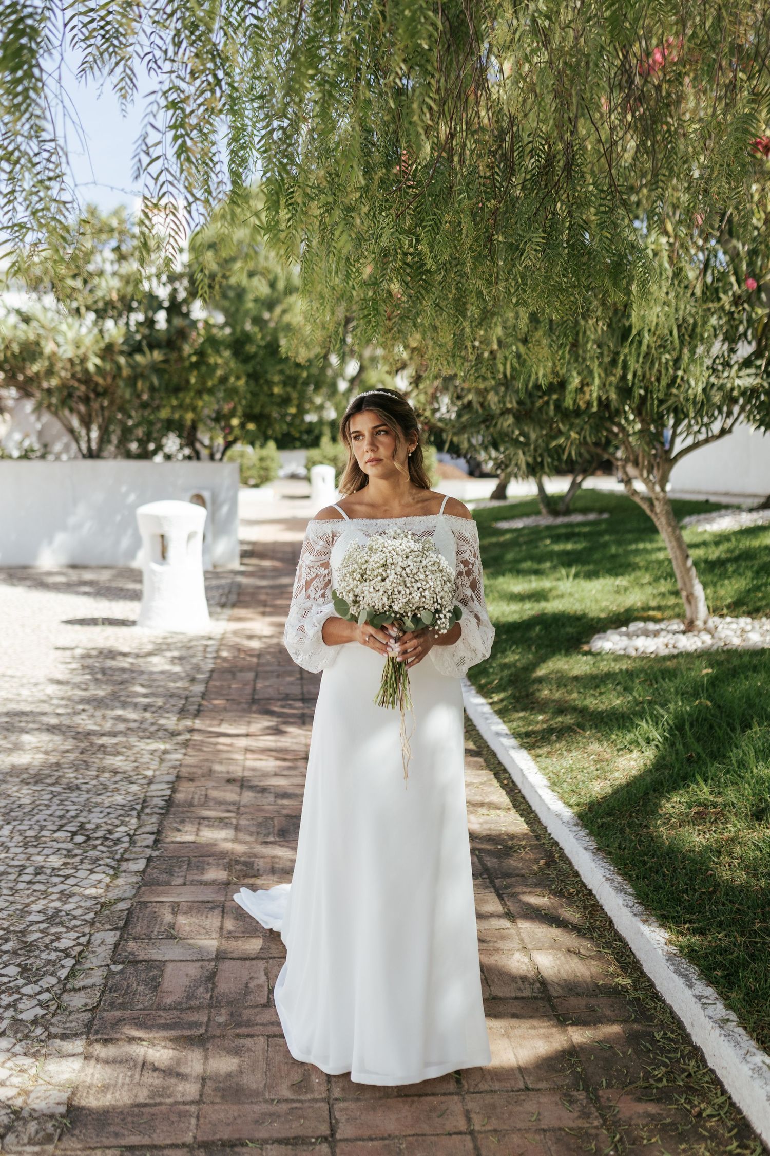 Elegant off-shoulder lace wedding dress with fit and flare silhouette shown on a garden path under weeping willow trees.