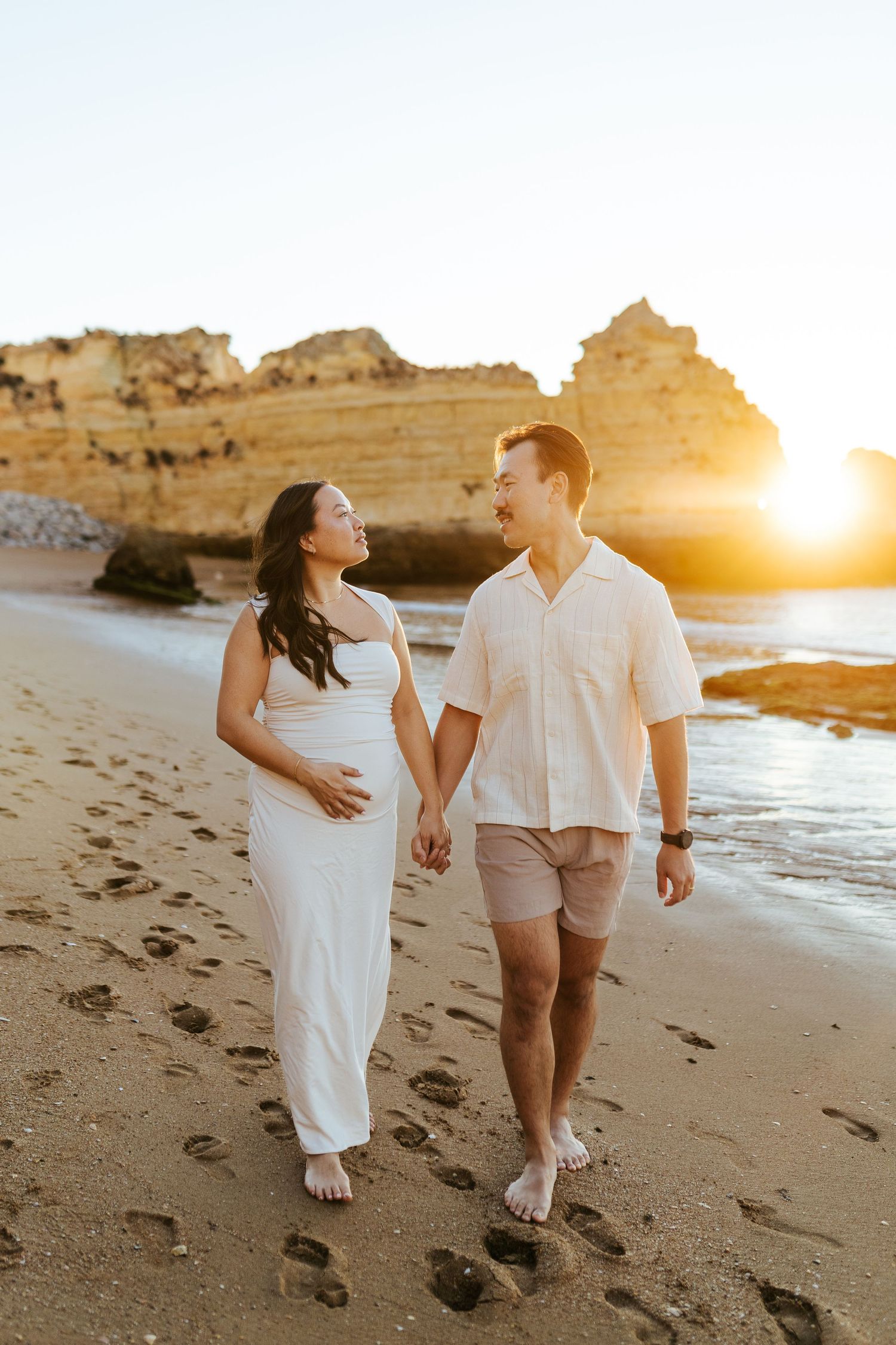 A couple strolls hand in hand on a sandy beach at sunset with rocky cliffs in the background.